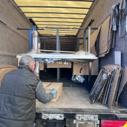 Twoja Przeprowadzka-Transport - Wnętrze ciężarówki z meblami biurowymi, pracownik w kamizelce wkłada karton. Kartony i zabezpieczenia na ścianach. Widok z tyłu pojazdu.