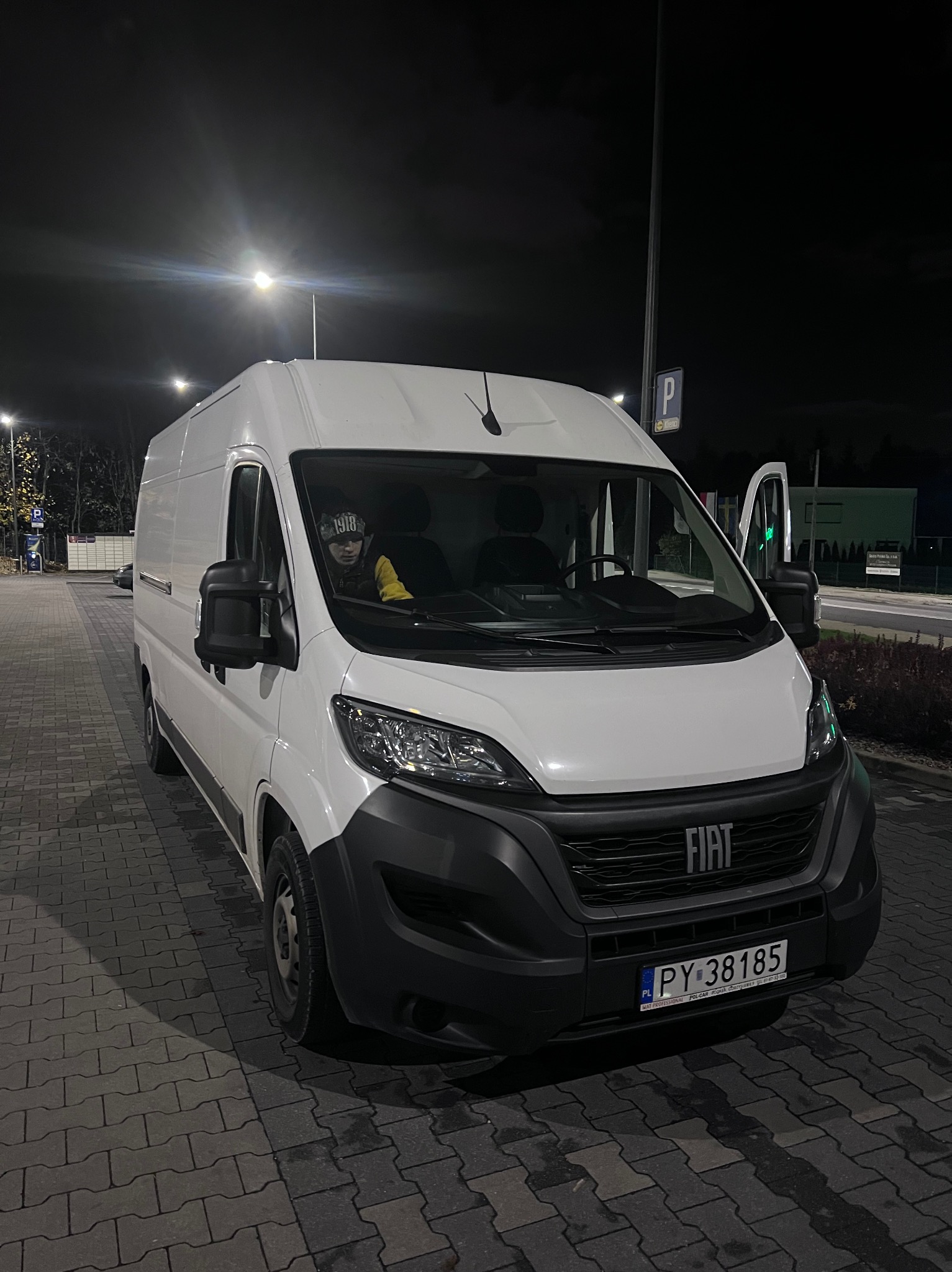 Biały furgon Fiat Ducato z otwartymi drzwiami pasażera, zaparkowany nocą na brukowanym parkingu, z widoczną osobą w środku i tablicą rejestracyjną PY 38185.