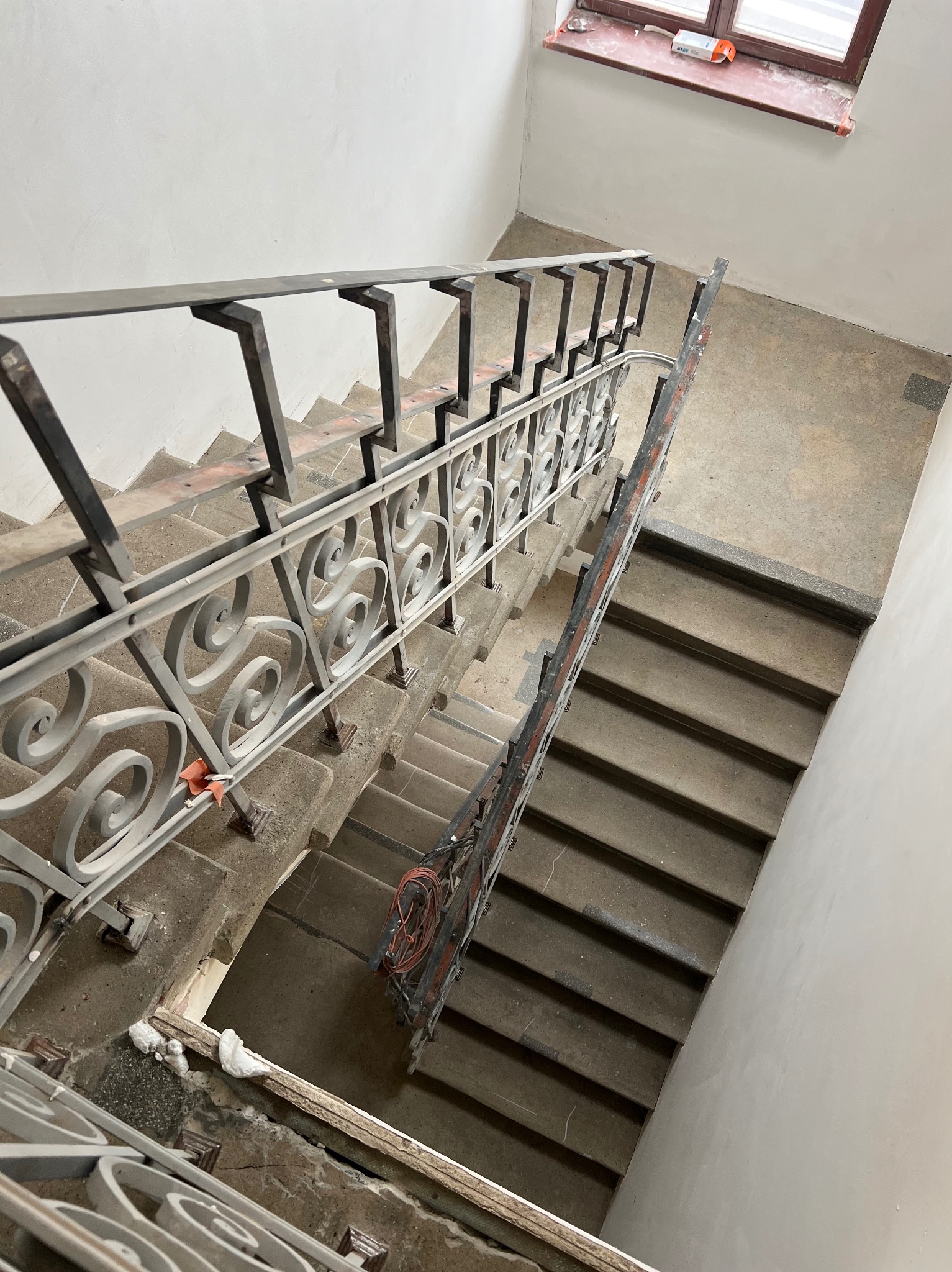 Widok z góry na betonowe schody z metalową balustradą o dekoracyjnym wzorze. Widoczne ślady prac remontowych, kabel i resztki materiałów budowlanych.