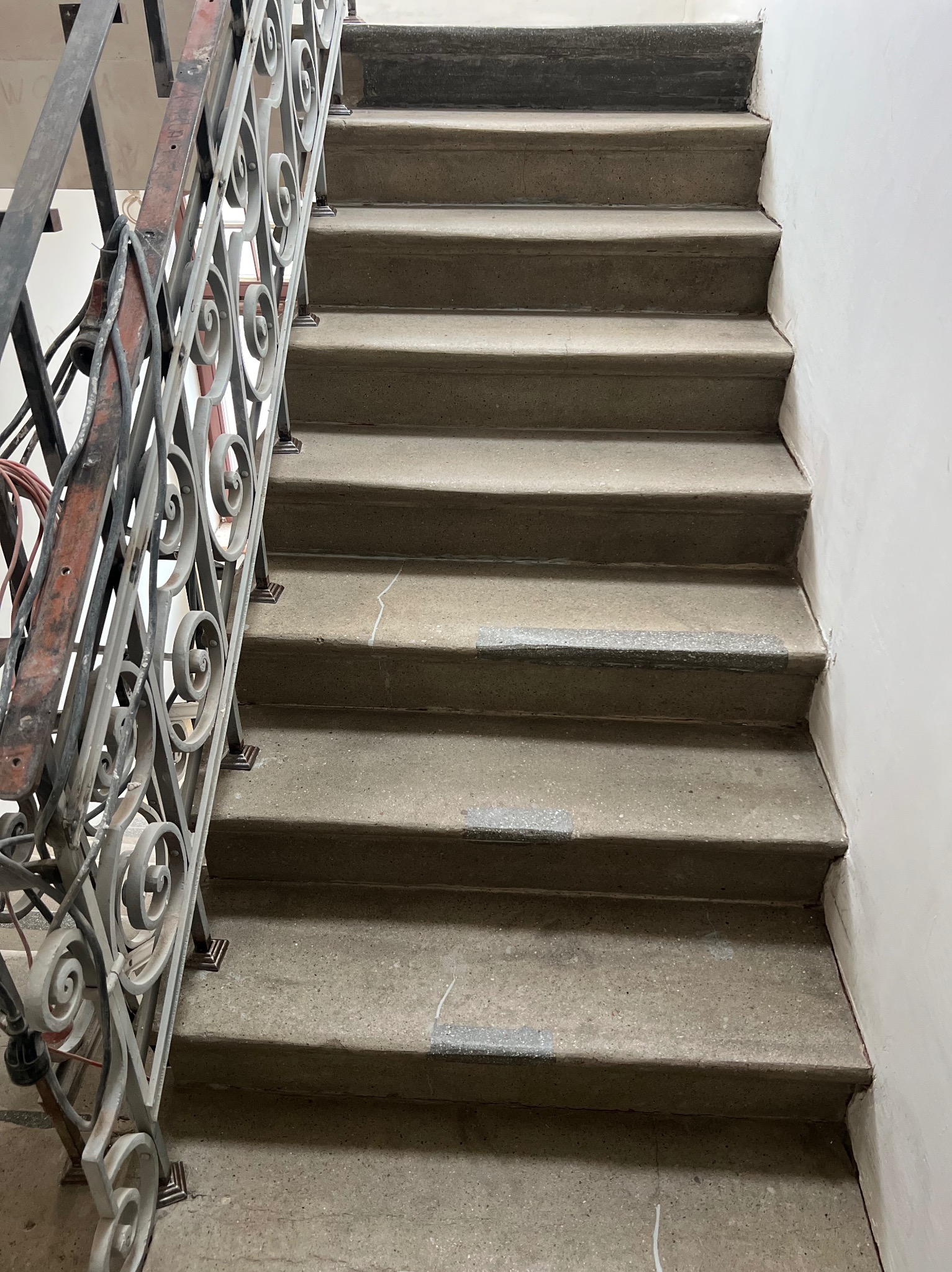 Betonowe schody z metalową, zdobioną balustradą. Widoczne ślady użytkowania i drobne uszkodzenia na stopniach. Ujęcie z dołu, podkreślające perspektywę w górę.