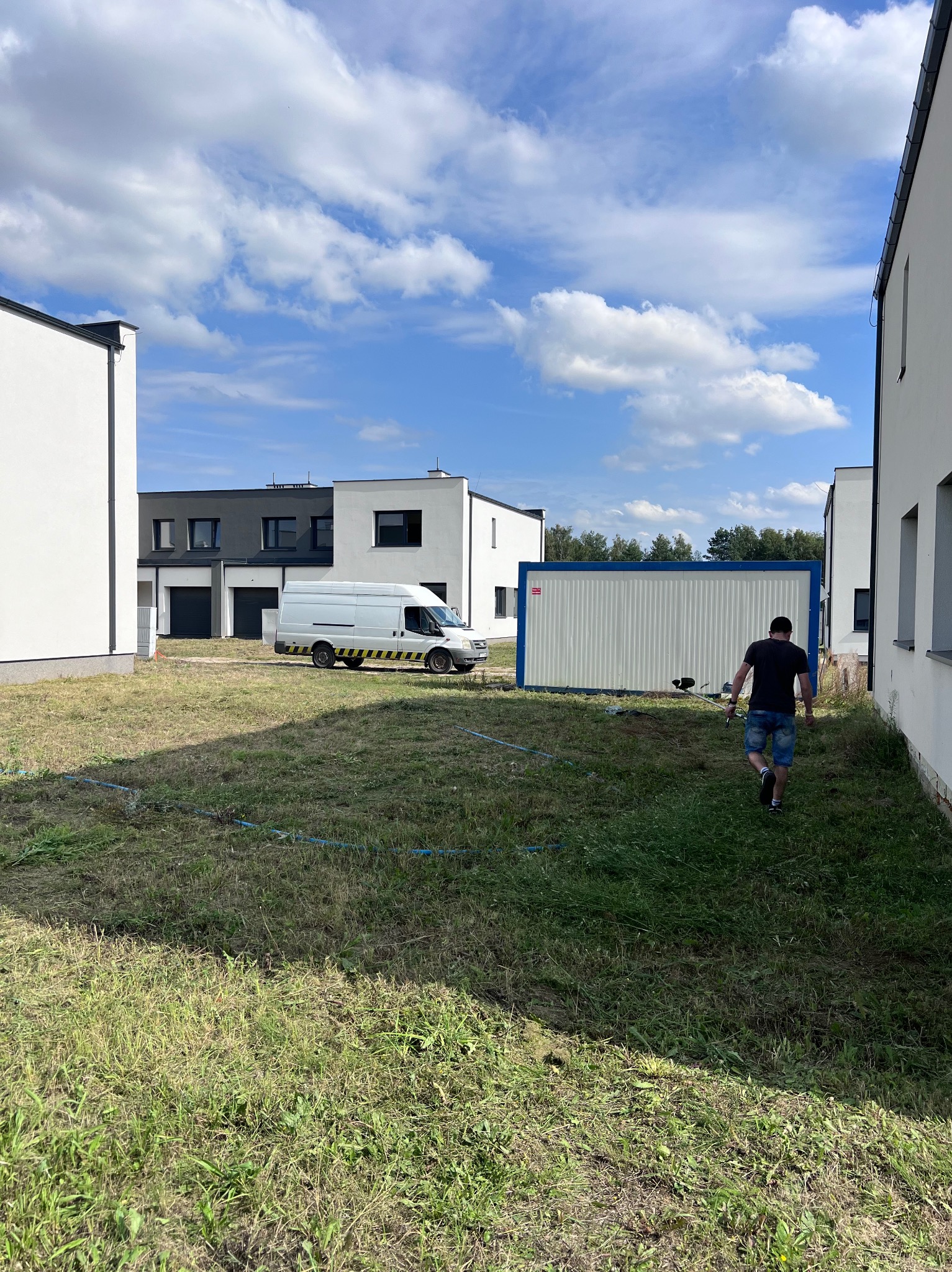 Przygotowanie terenu: mężczyzna z kosiarką idzie w stronę kontenera budowlanego na zarośniętej działce między nowoczesnymi domami. Biały bus z żółtymi pasami w tle.