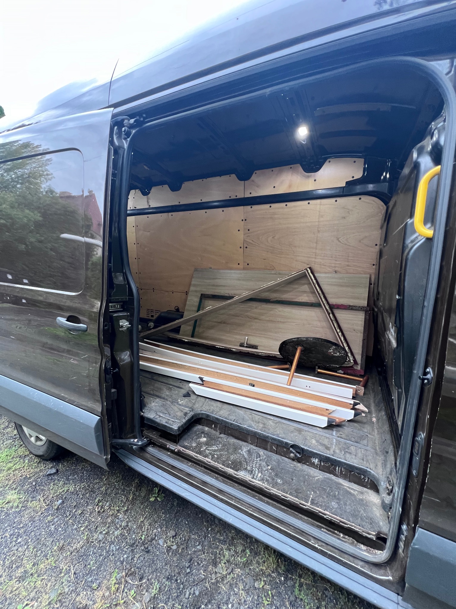 Otwarty brązowy van z drewnianymi elementami i odpadami w środku, prawdopodobnie po demontażu lub opróżnianiu. Widoczne elementy stolarki i fragmenty mebli.