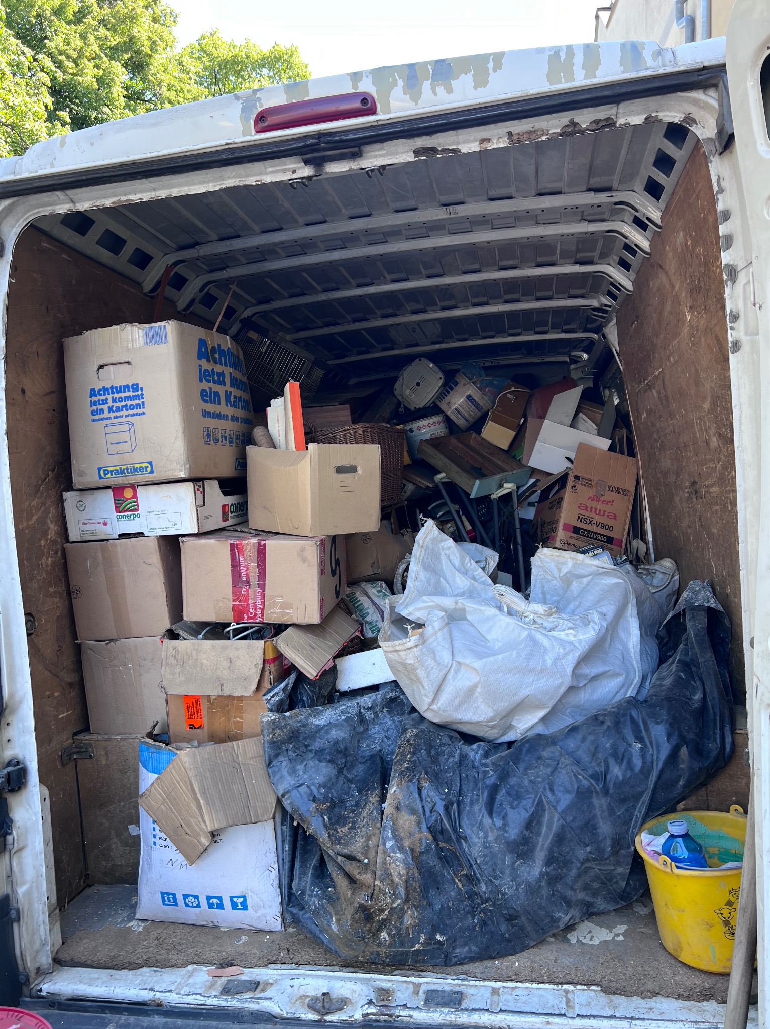 Wnętrze furgonetki załadowane kartonami, workami i różnymi przedmiotami, przygotowane do transportu. Widoczny żółty kubeł z płynem.