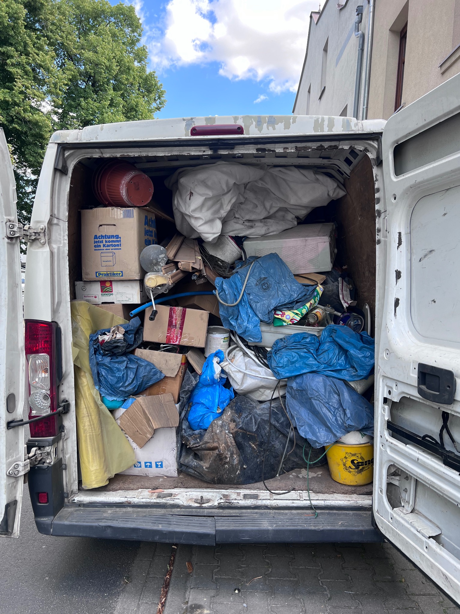 Otwarty bagażnik białego vana wypełniony kartonami, workami z odpadami i różnymi przedmiotami. Widoczne elementy wyposażenia wnętrza i fragmenty karoserii pojazdu.