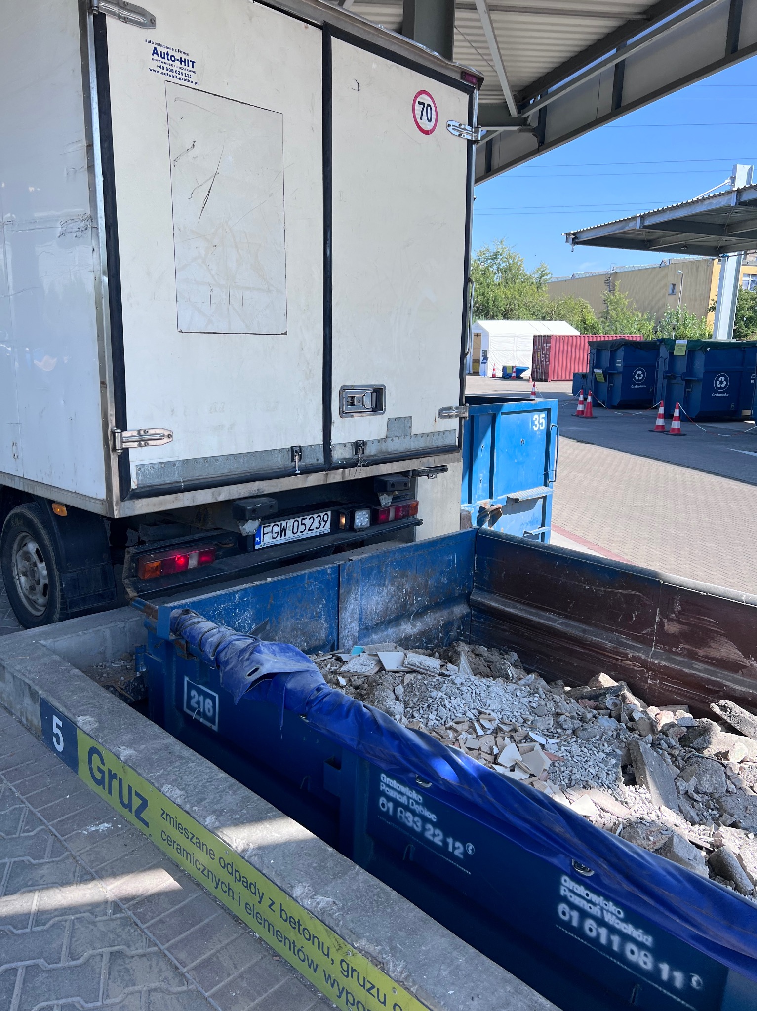 Ciężarówka z otwartymi drzwiami załadunkowymi, tyłem do kontenera wypełnionego gruzem i odpadami budowlanymi na terenie punktu recyklingu.