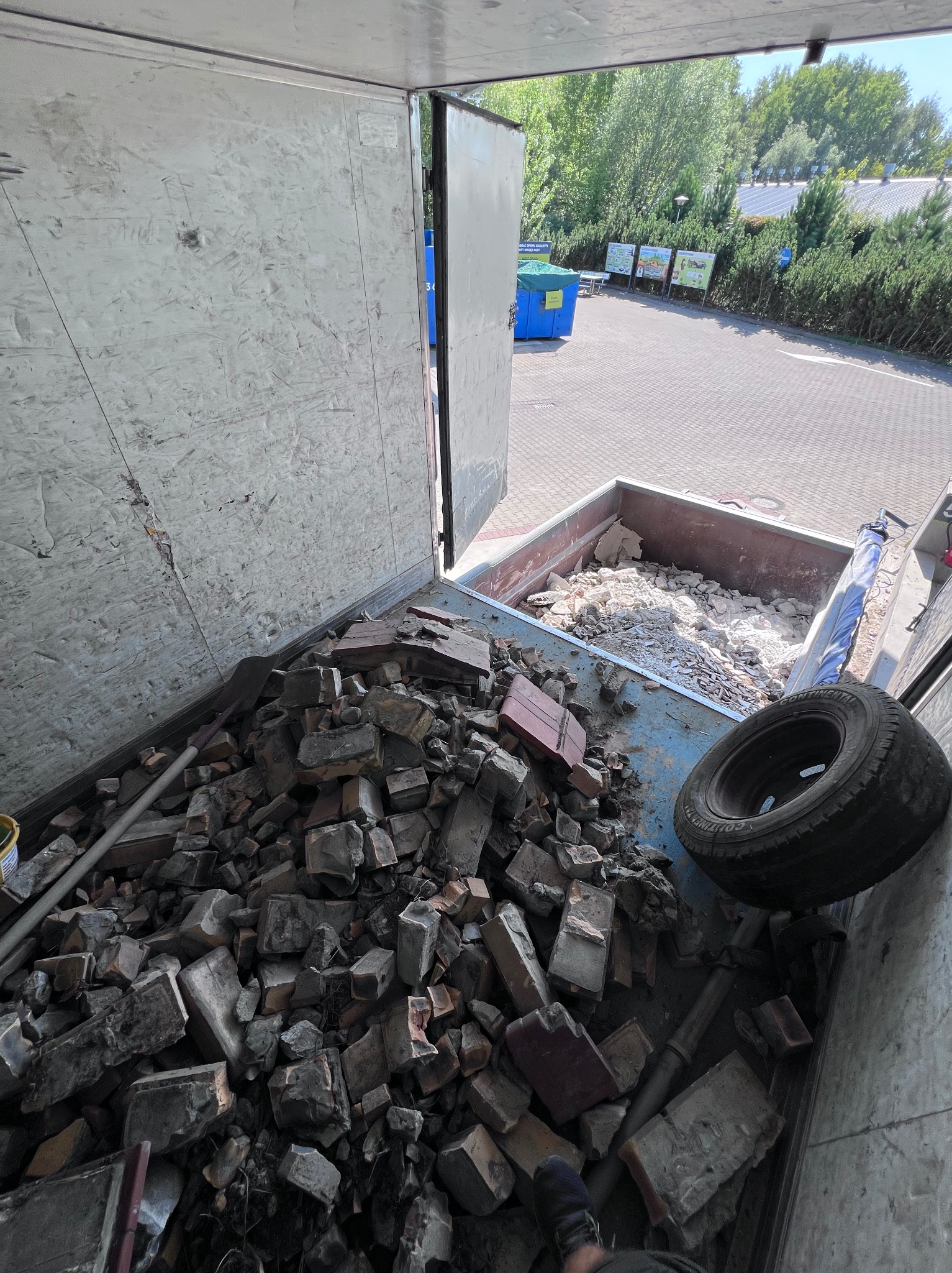 Gruz ceglany i gruz betonowy w kontenerze, widok z góry, Poznań. Widoczne narzędzia i opona. Kontener otwarty, w tle zieleń i budynki.