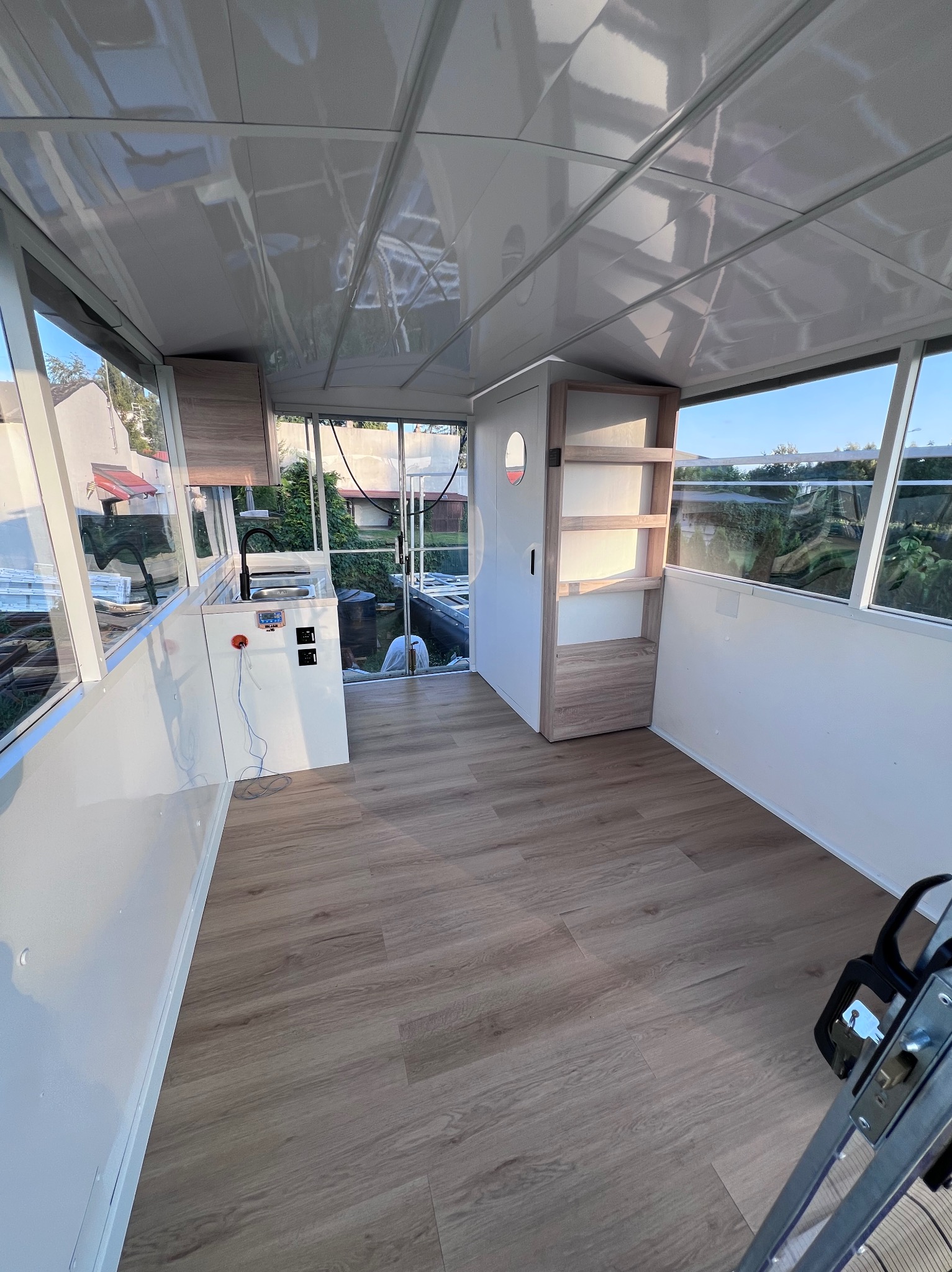 Jasne wnętrze modułowego domku letniskowego z laminowaną podłogą, białymi ścianami i dużymi oknami. Widoczny aneks kuchenny i regał. Na zewnątrz zieleń.