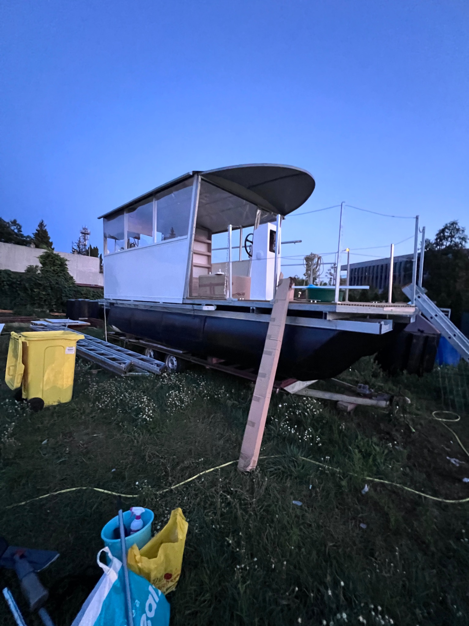 Nietypowy, częściowo zadaszony houseboat w trakcie budowy na trawniku, otoczony narzędziami i materiałami budowlanymi. Widoczne elementy konstrukcyjne i robocze otoczenie.