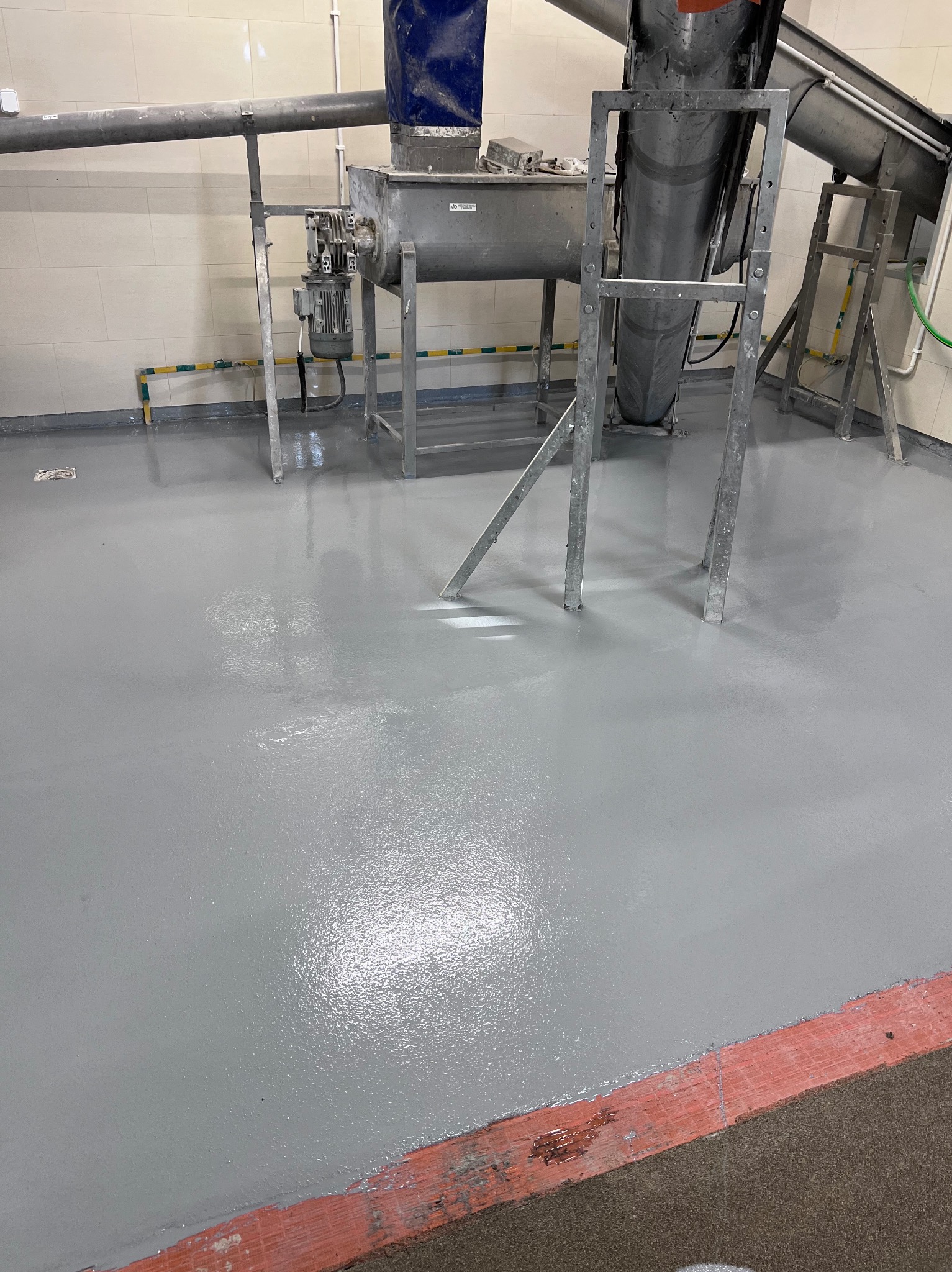 Błyszcząca, szara posadzka przemysłowa z metalowymi konstrukcjami maszyn w tle. Widoczne detale wykończenia przy ścianach. Gładka powierzchnia, jasne oświetlenie.