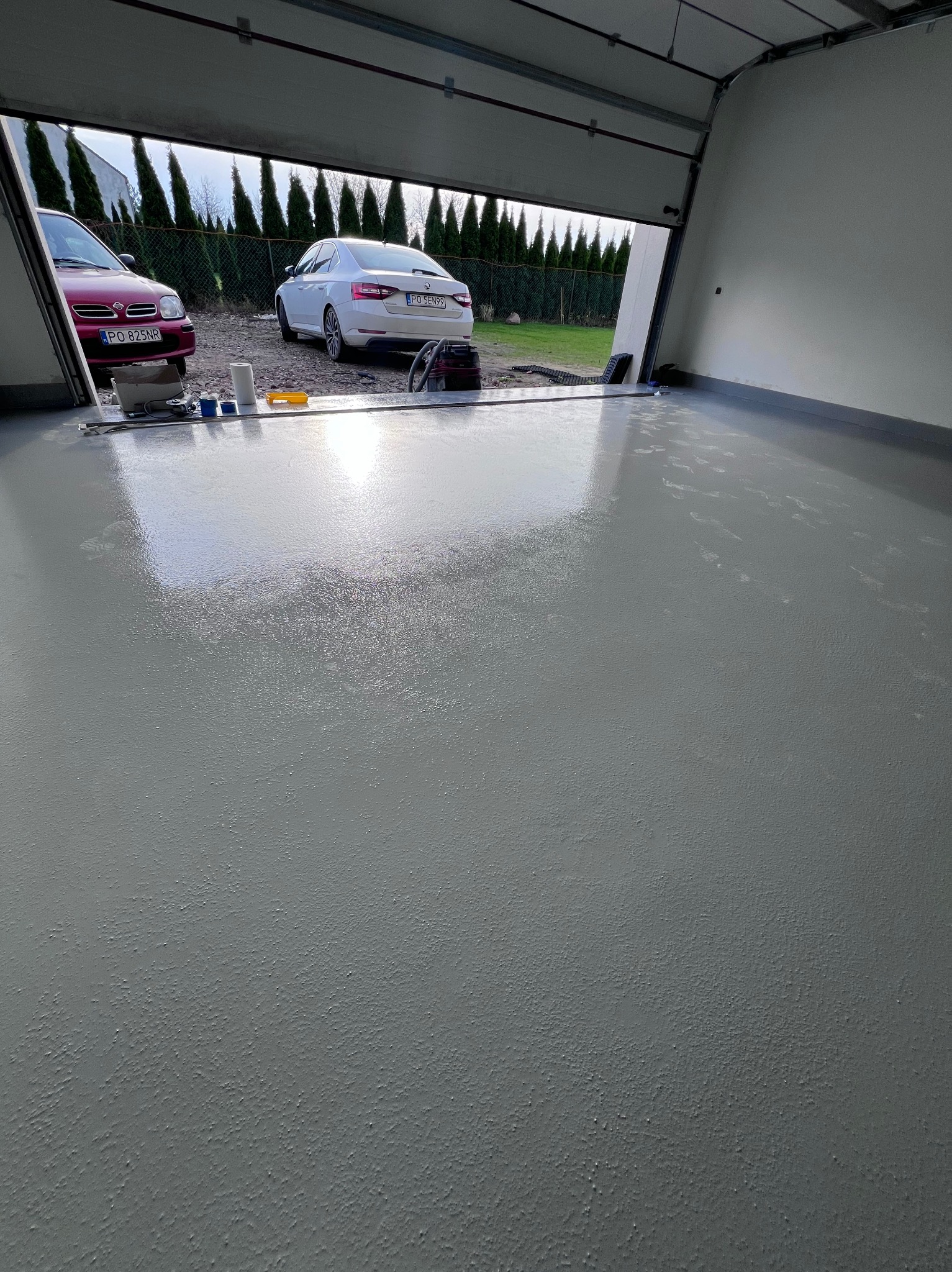 Świeżo wylana, szara posadzka w garażu z otwartą bramą, przez którą widać dwa samochody i zieleń. Na posadzce widoczne ślady i narzędzia malarskie.