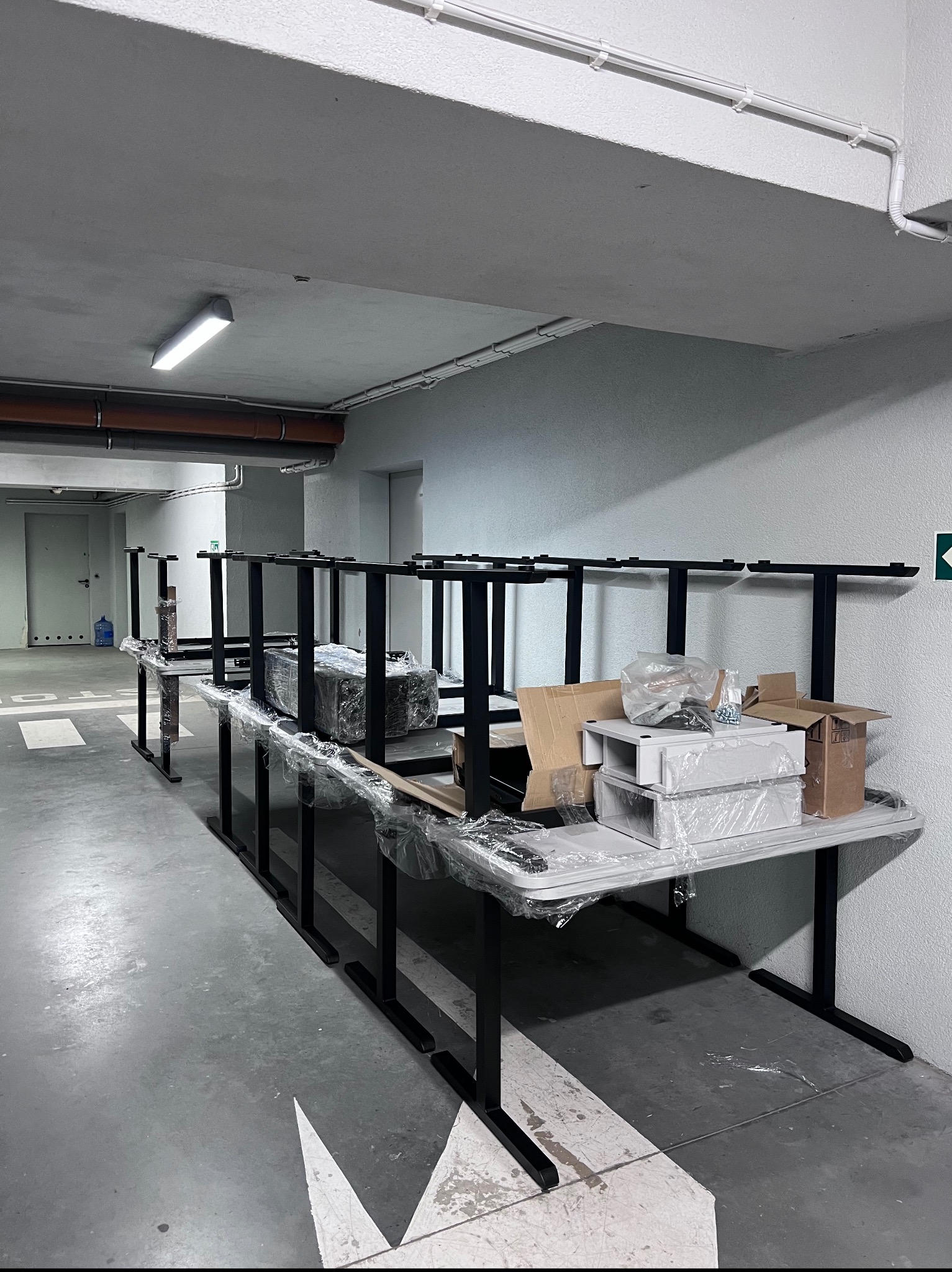 Rząd rozłożonych, ale jeszcze zapakowanych stołów z czarnymi nogami w szarym garażu podziemnym. Na blatach kartony i elementy montażowe w folii.
