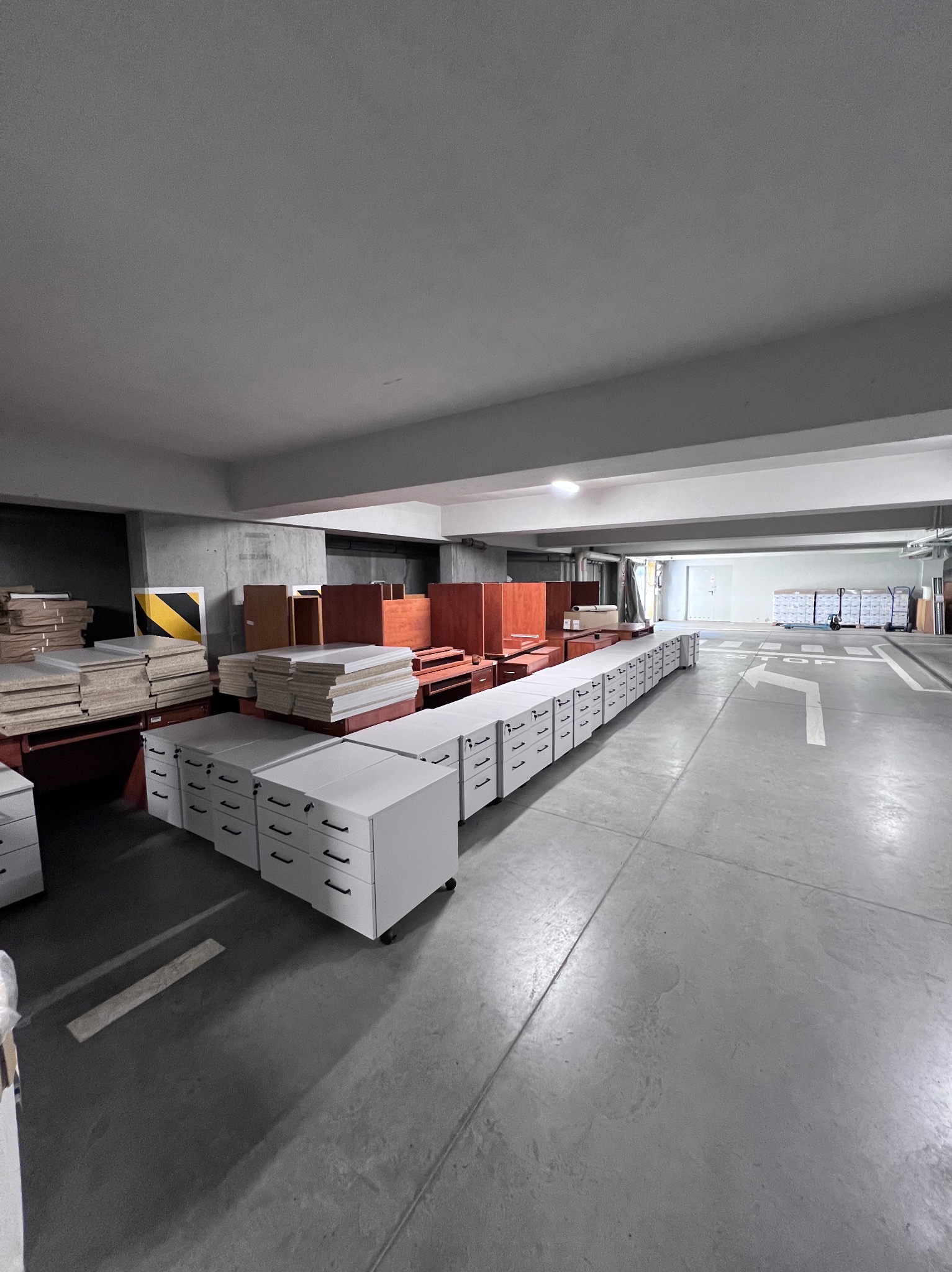 Magazyn z rzędem białych szafek na kółkach i stertą płyt meblowych, gotowych do transportu. Widok z perspektywy szerokokątnej, ukazujący przestrzeń i przygotowanie do przeprowadzki.