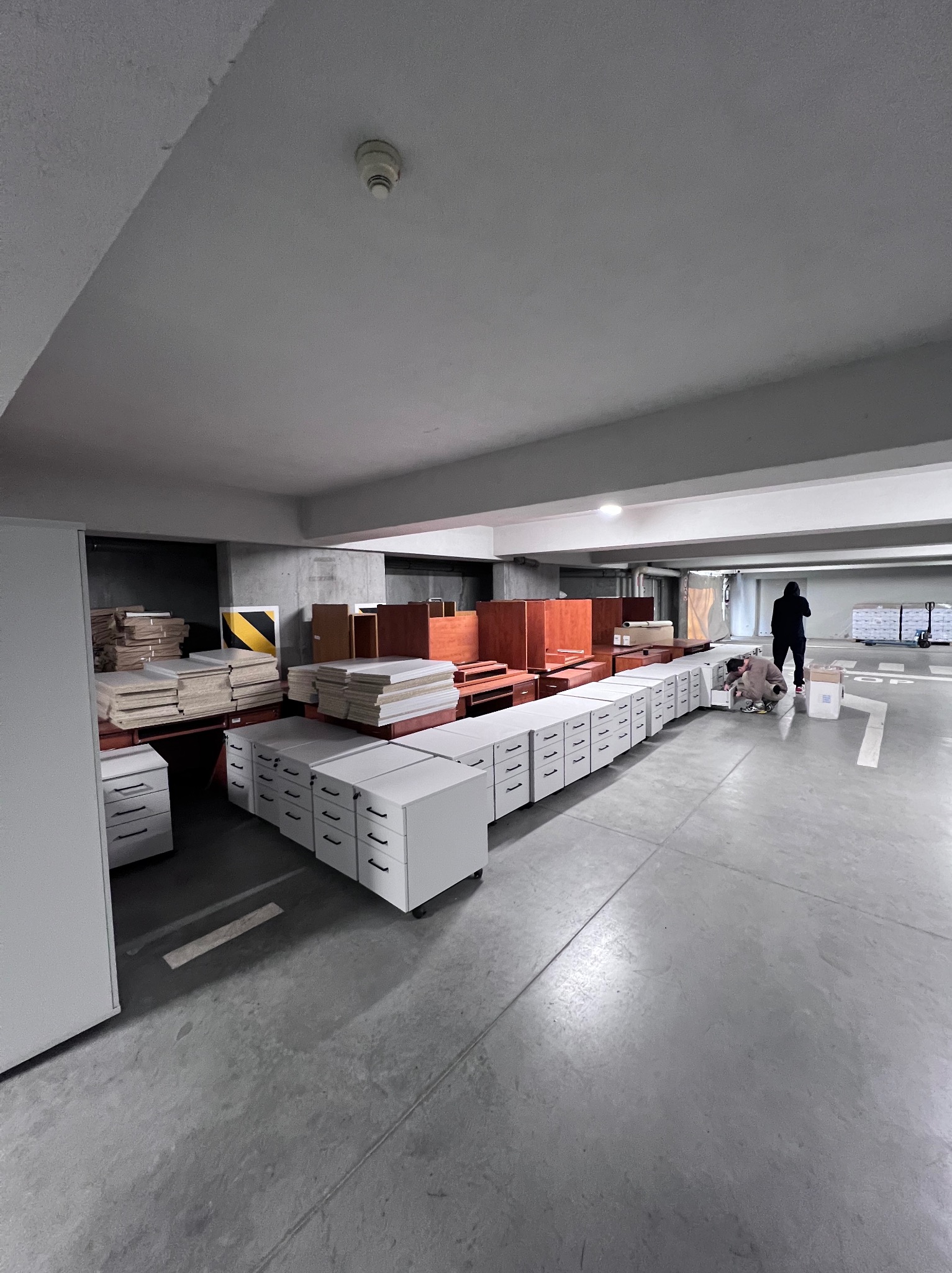 Montaż mebli biurowych w garażu podziemnym. Rząd białych szafek z szufladami, elementy drewniane, pracownik w trakcie montażu. Oświetlenie sufitowe, betonowa podłoga.