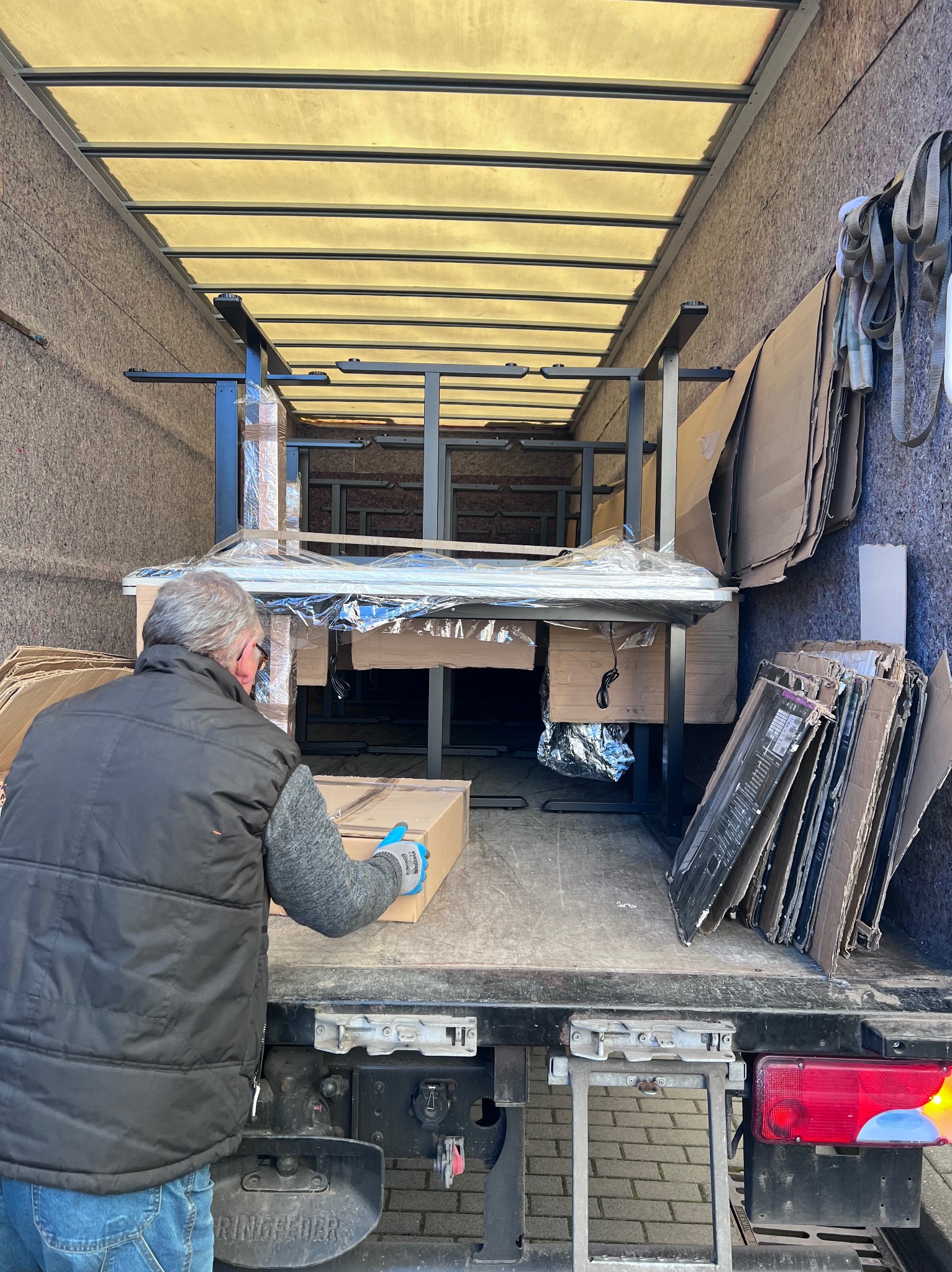 Wnętrze ciężarówki z meblami biurowymi, pracownik w kamizelce wkłada karton. Kartony i zabezpieczenia na ścianach. Widok z tyłu pojazdu.