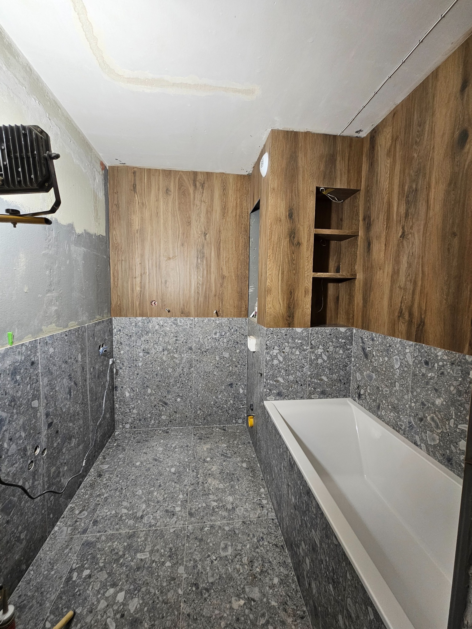 Remont łazienki w trakcie: szare płytki na podłodze i ścianach do połowy wysokości, drewnopodobne panele na górze. Widoczna wanna i oświetlenie robocze.