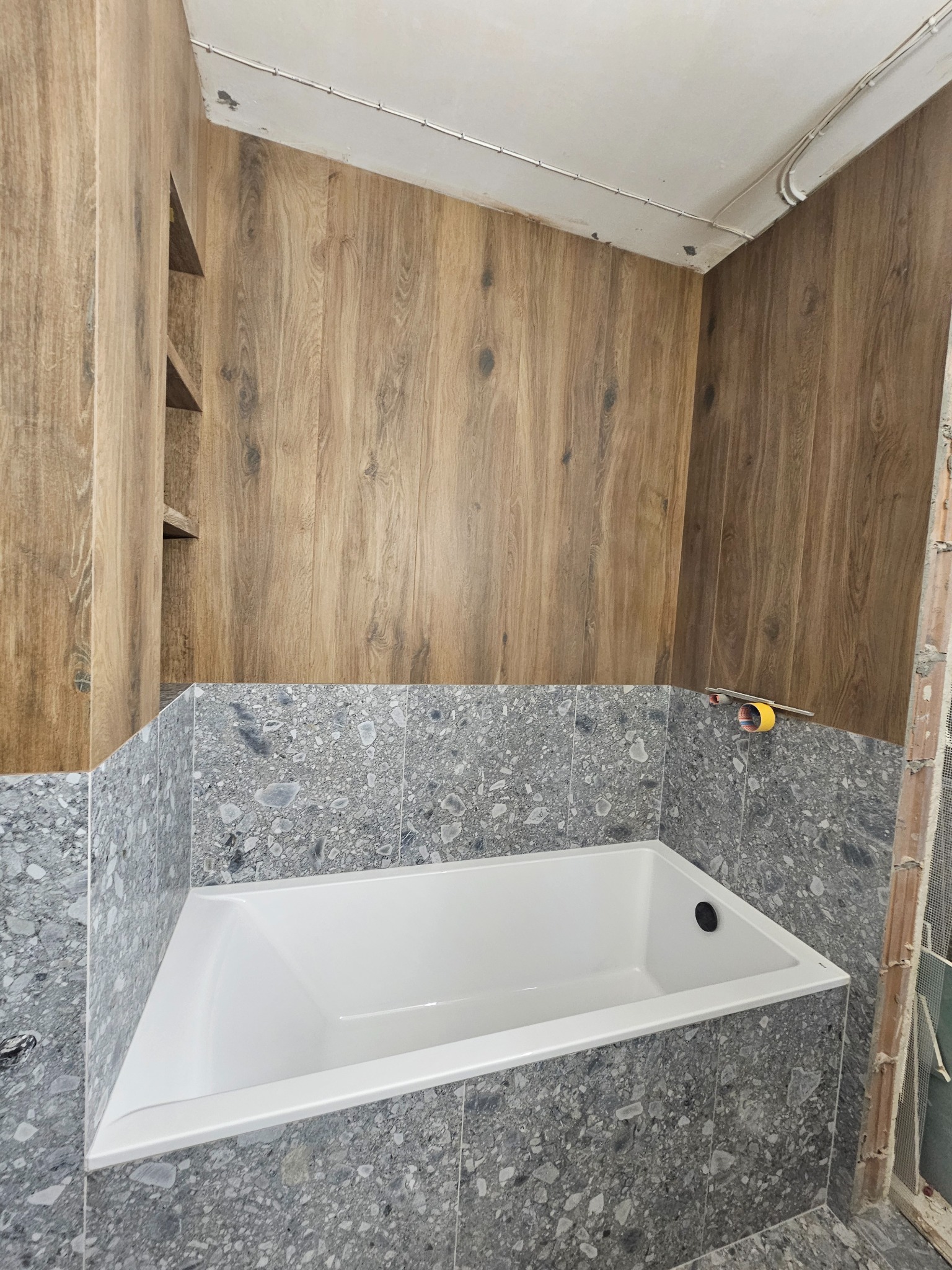 Wykończona łazienka z wanną obudowaną szarą mozaiką, drewnopodobne panele na ścianach i półki. Widoczne narzędzia glazurnicze.