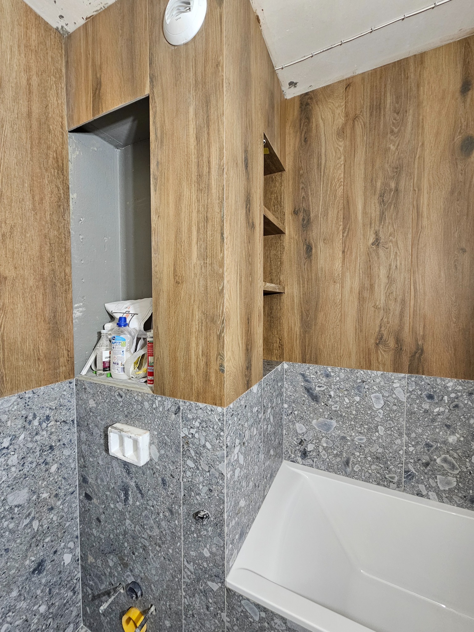 Wykończona łazienka w Krakowie: szare płytki z wzorem lastryko na ścianach i obudowie wanny, drewnopodobne panele na górze, półki i wnęka.