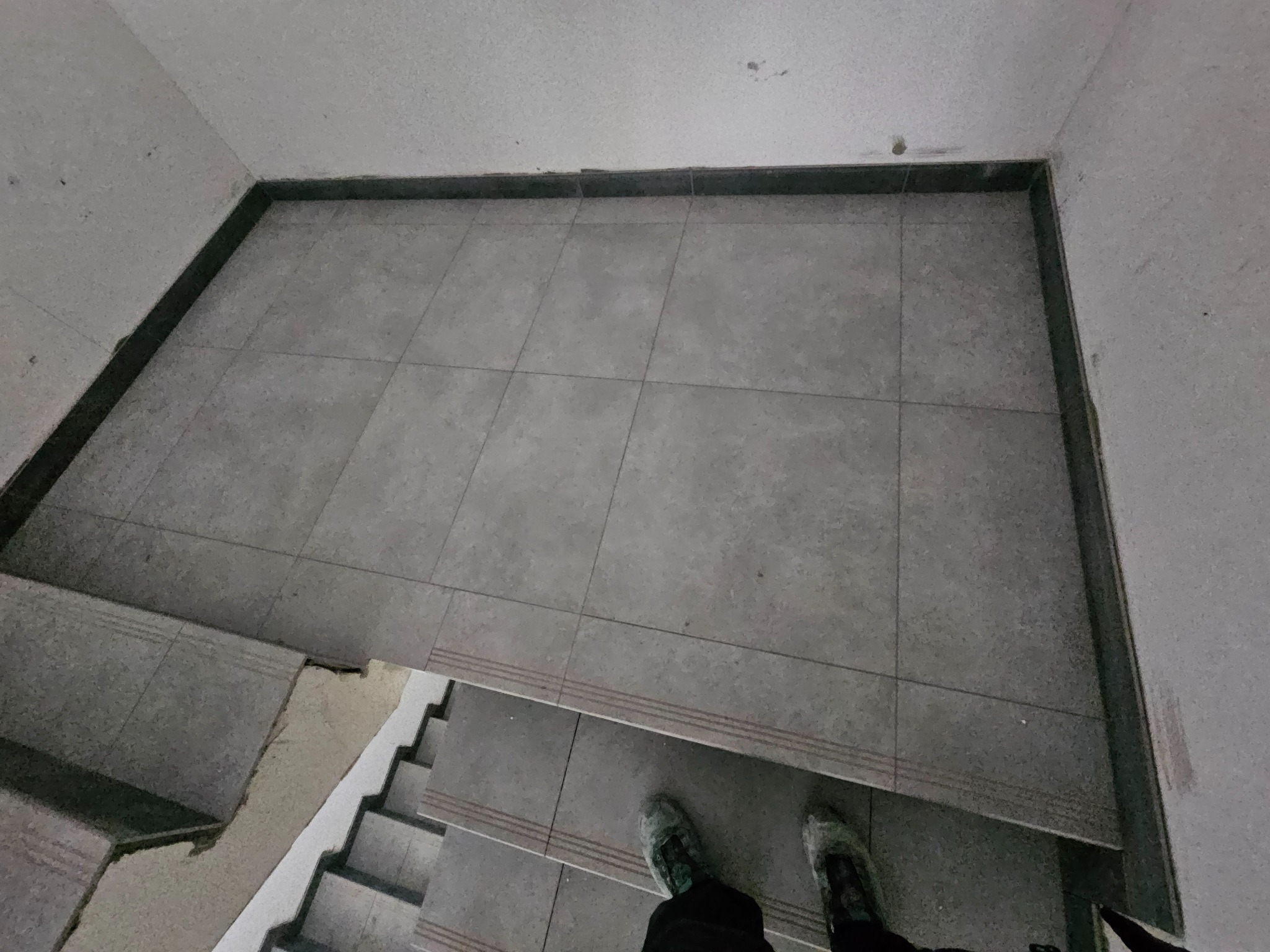 Szare płytki podłogowe w korytarzu, widok z góry na schody. Fragment butów w dolnej części kadru. Minimalistyczne wykończenie wnętrza.