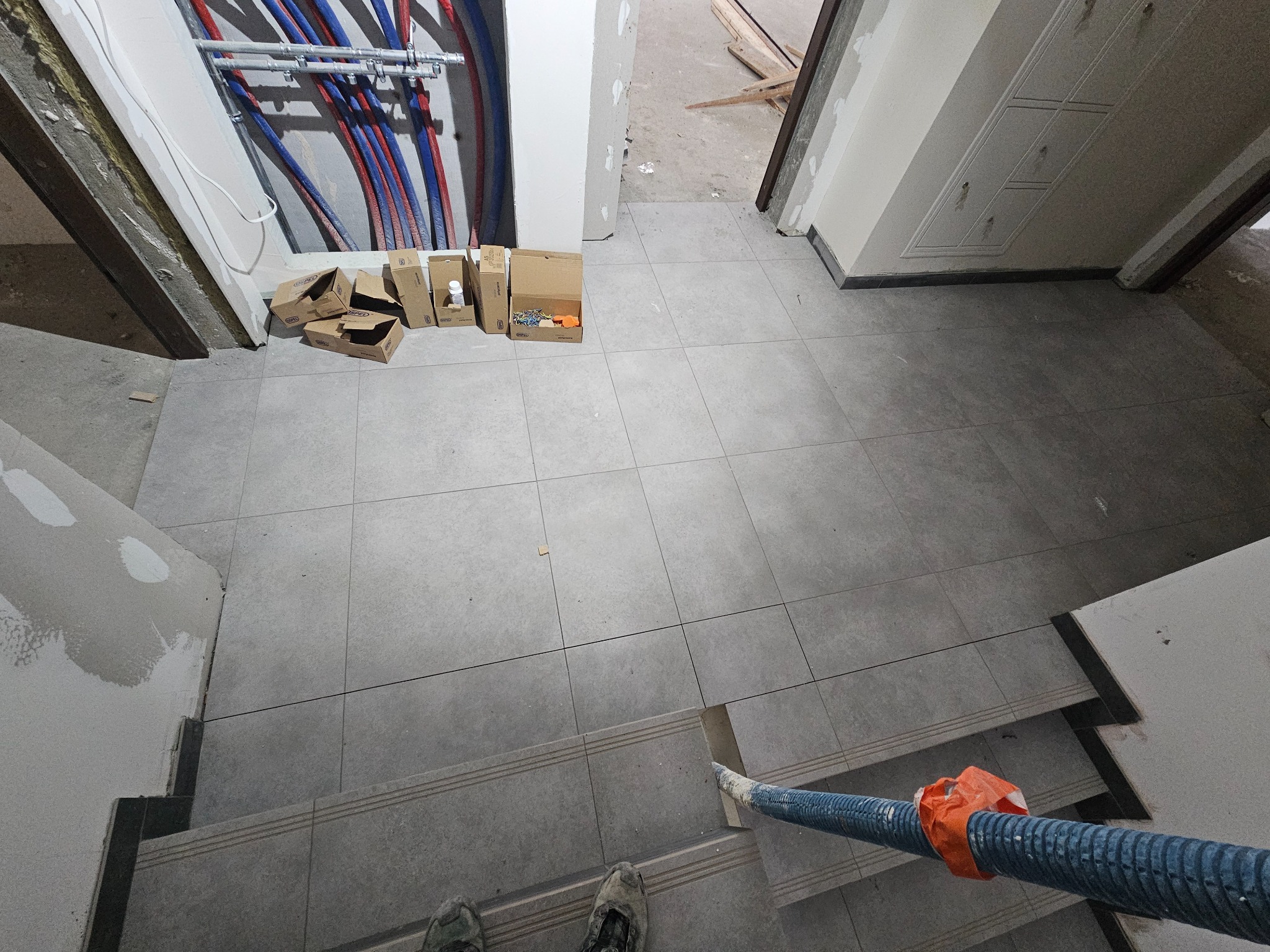 Szare płytki podłogowe ułożone na schodach i podłodze, widoczne rury instalacyjne przy ścianie, kartony z akcesoriami i odkurzacz przemysłowy. Ukończony etap prac wykończeniowych.