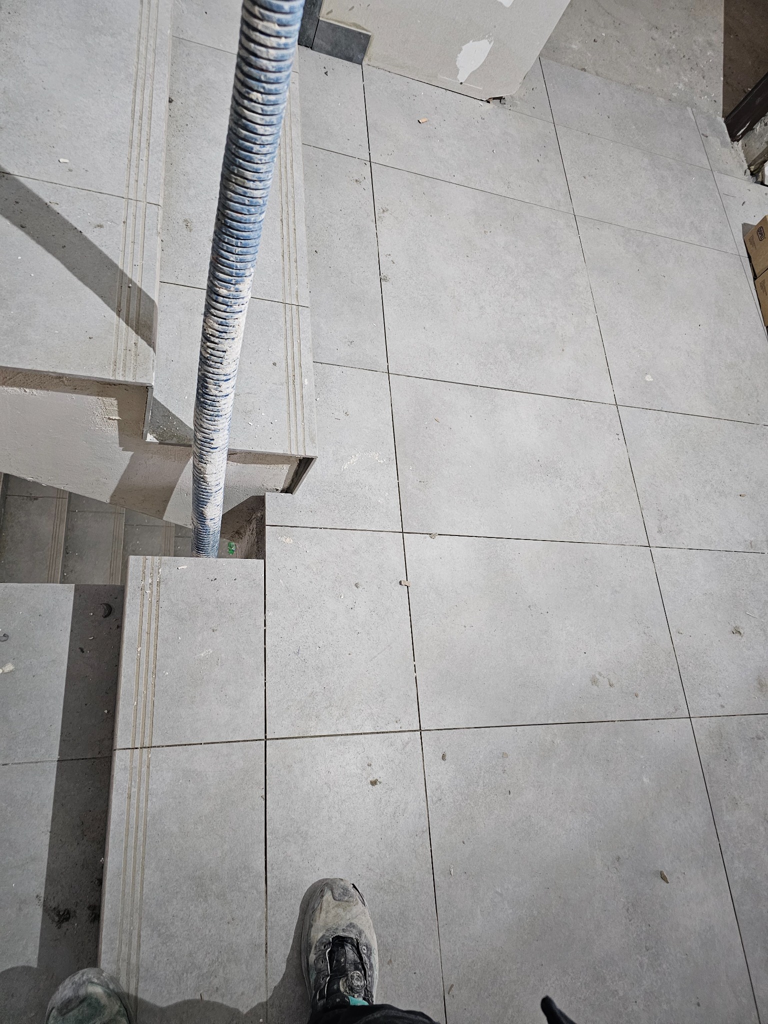 Ułożone szare płytki podłogowe i schodowe z niebieskim wężem odkurzacza. Widok z góry na fragment podłogi i schodów w trakcie prac wykończeniowych.