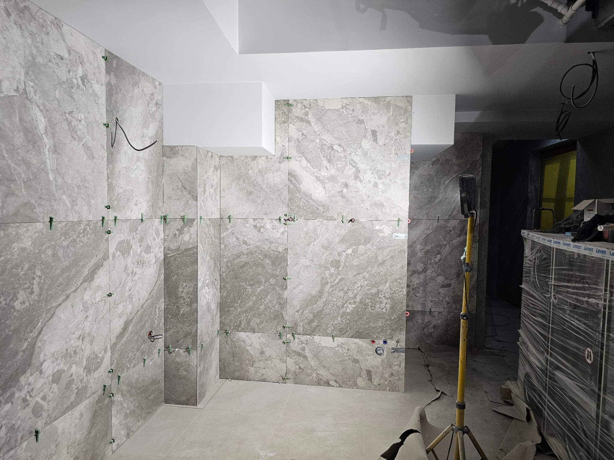 Ściany i podłoga w trakcie układania płytek imitujących marmur. Widoczne krzyżyki dystansowe i poziomica laserowa. Oświetlenie halogenowe.