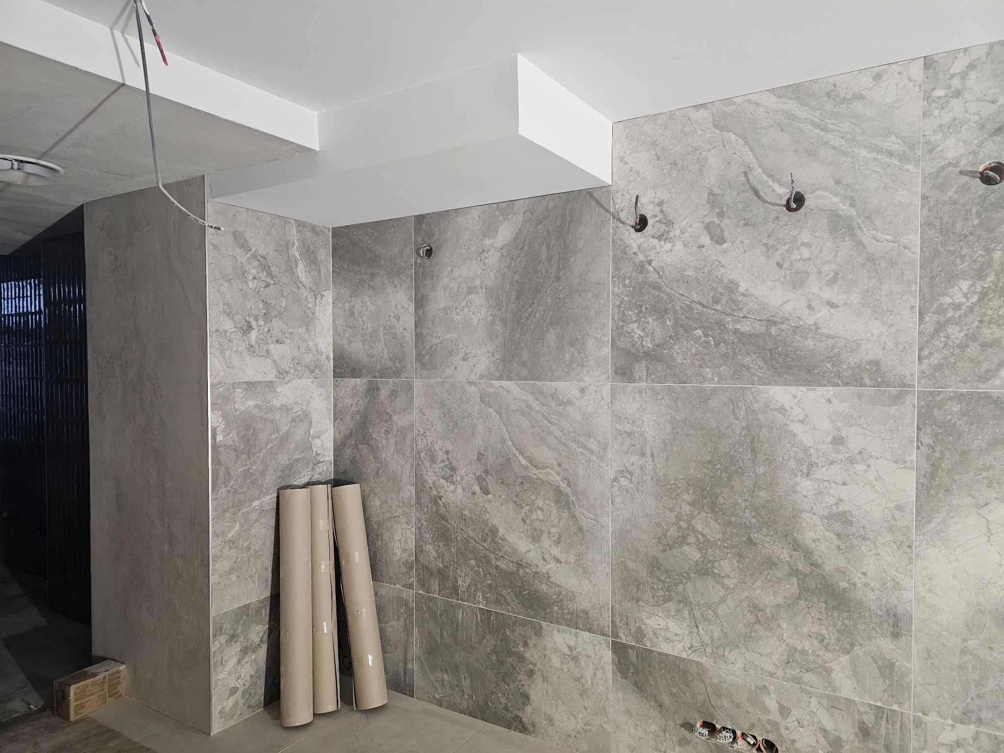 Wykończona łazienka z szarymi płytkami imitującymi marmur, widoczne przygotowane punkty oświetleniowe i rury z nowymi płytkami w rolkach.