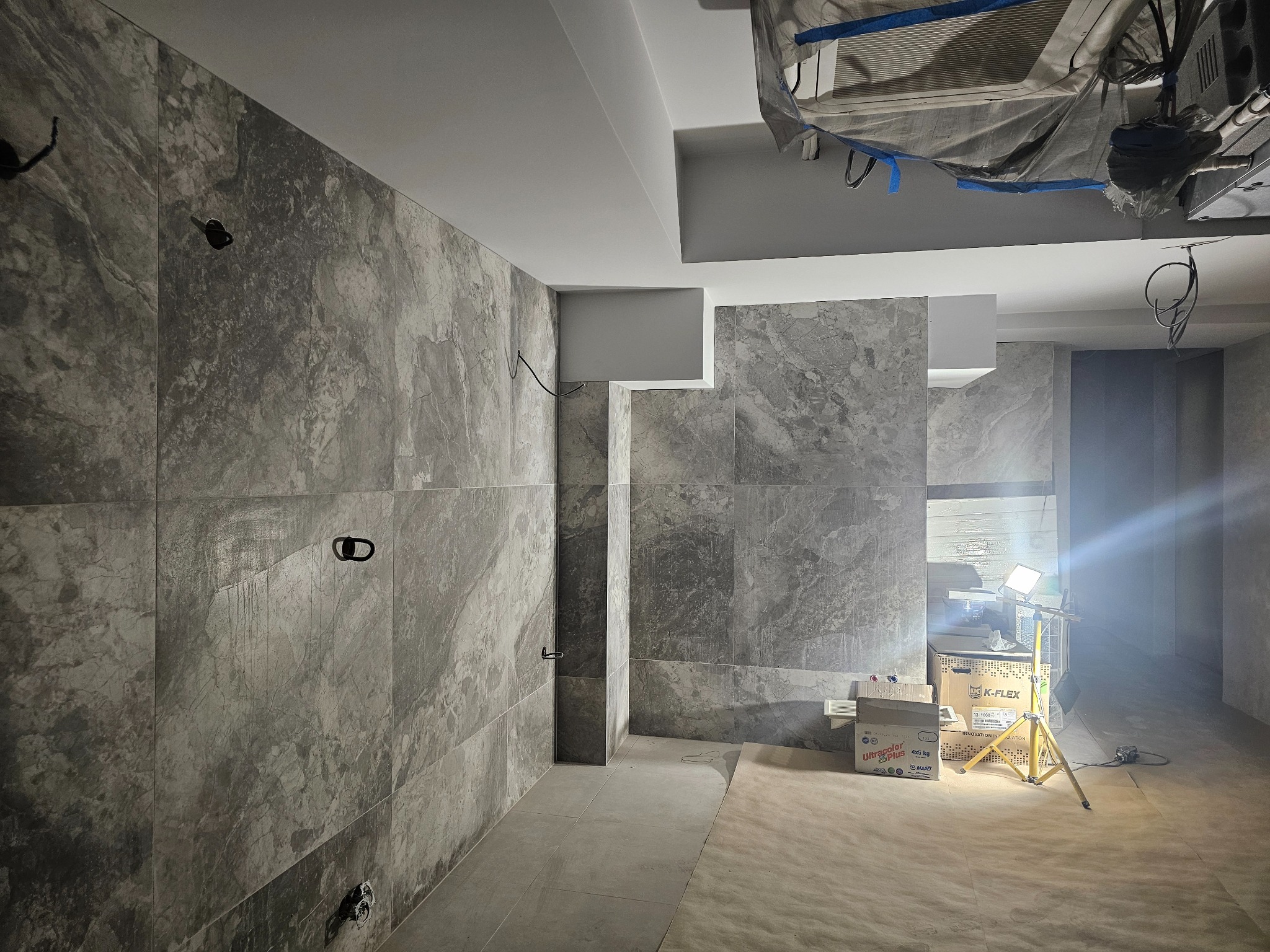 Układanie płytek imitujących marmur w łazience, widoczne puszki elektryczne i lampa budowlana. Szare płytki na ścianach i podłodze, w tle kartony z klejem do płytek.