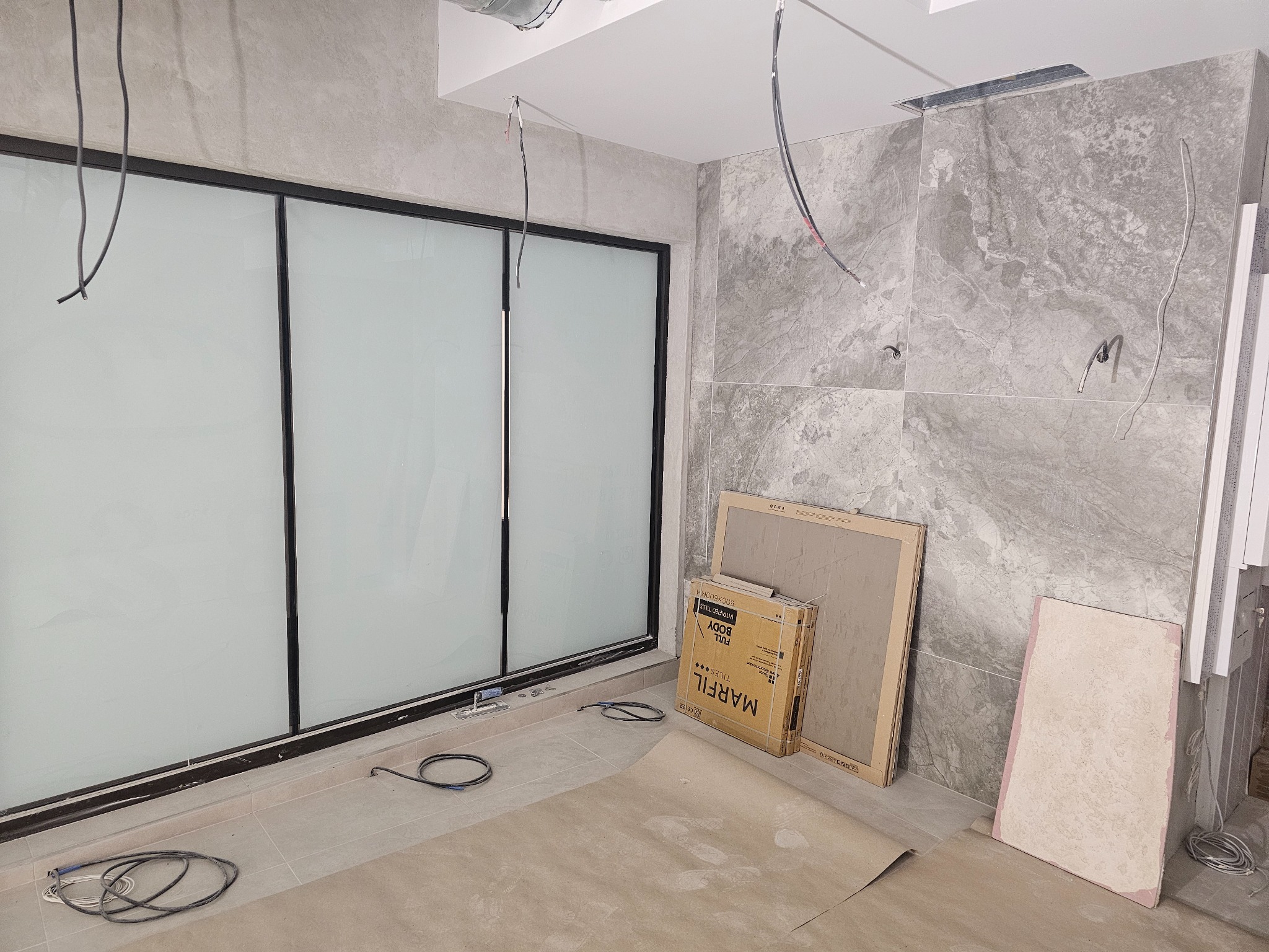 Wnętrze w trakcie prac wykończeniowych: szare płytki imitujące marmur na ścianach, kartony z płytkami Marfil, zabezpieczona podłoga. Widoczne przewody elektryczne.