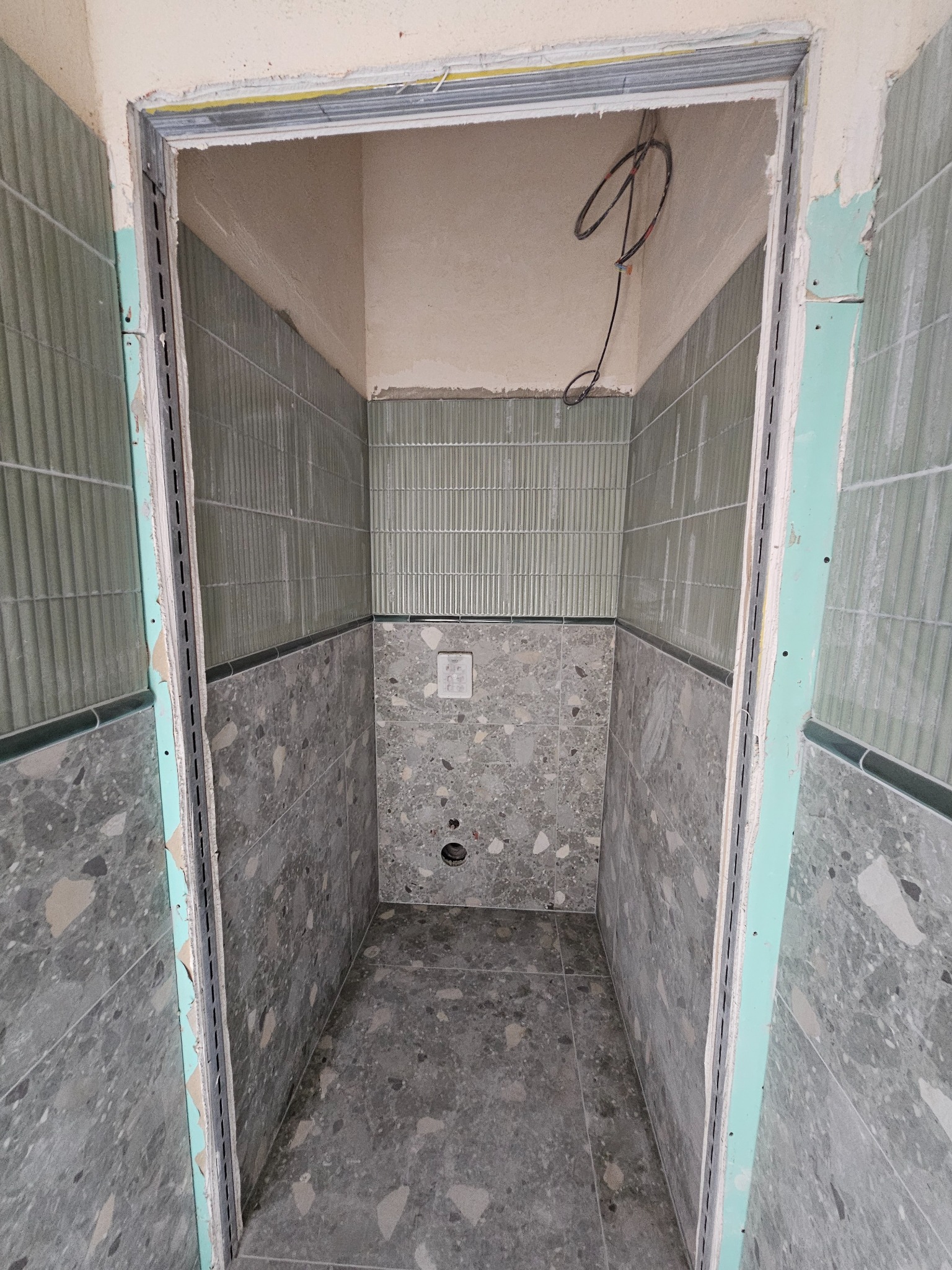 Wnętrze łazienki w trakcie remontu. Widoczne płytki w dwóch odcieniach szarości, pionowe żłobienia na górze i wzór lastryko na dole. Metalowe profile przy drzwiach.