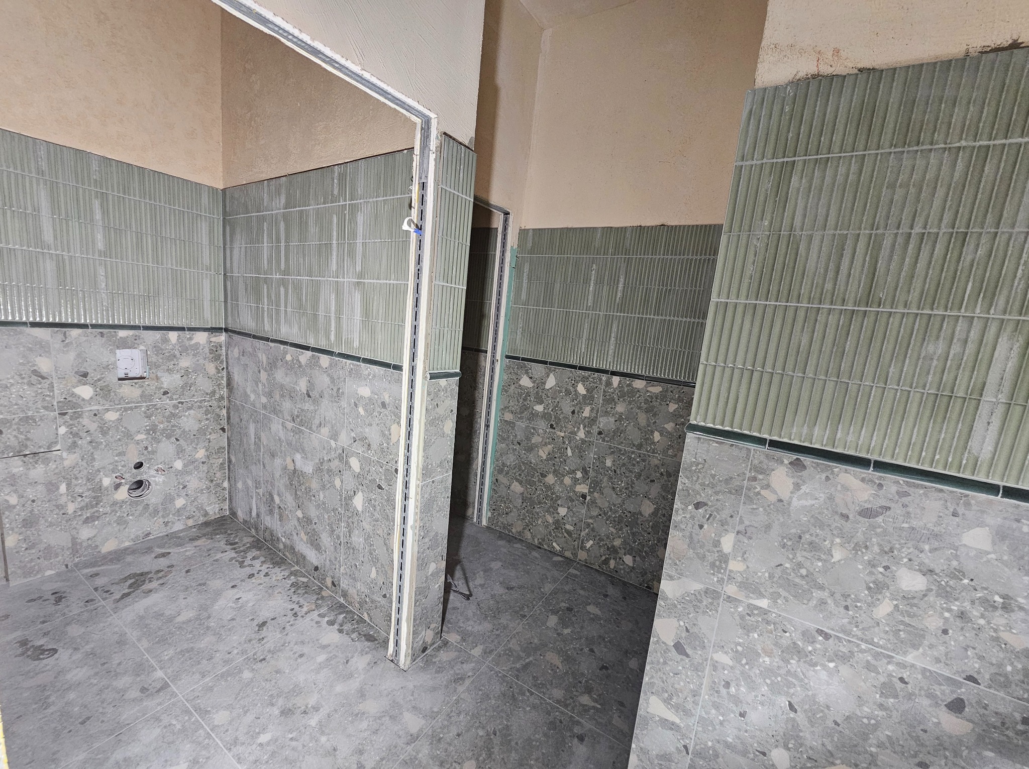 Wykończona łazienka z szarymi płytkami lastryko na podłodze i ścianach do połowy wysokości, powyżej zielone, ryflowane płytki. Widoczne ościeżnice drzwi.