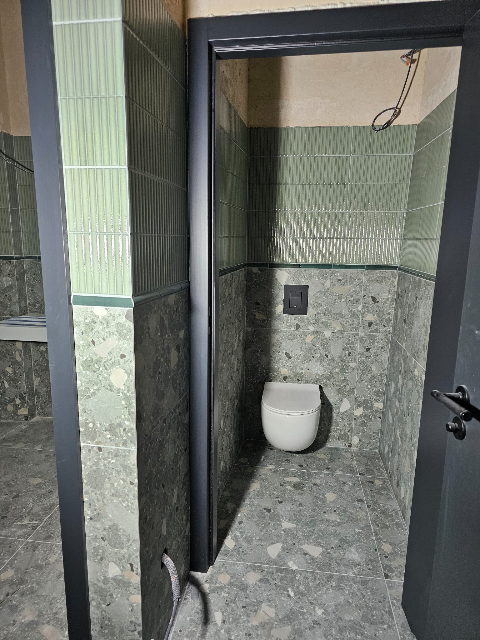 Wykończona łazienka z zielonymi, żłobionymi płytkami na górze i terazzo na dole. Widoczna toaleta i fragment umywalki. Ciemna rama drzwi.