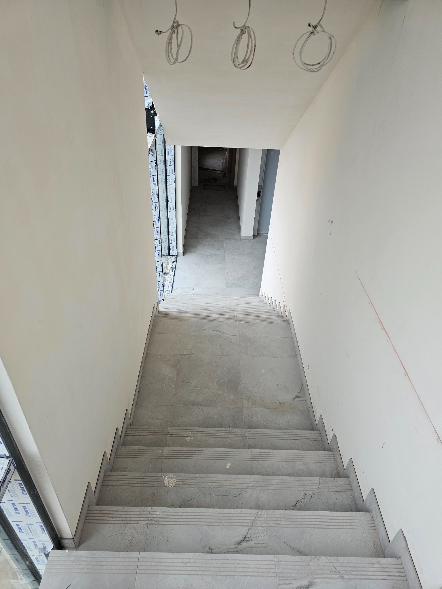 Wykończone schody z szarych płytek imitujących marmur, widok z góry. Białe ściany i sufit, widoczne kable oświetleniowe. Nowoczesne wykończenie wnętrza.