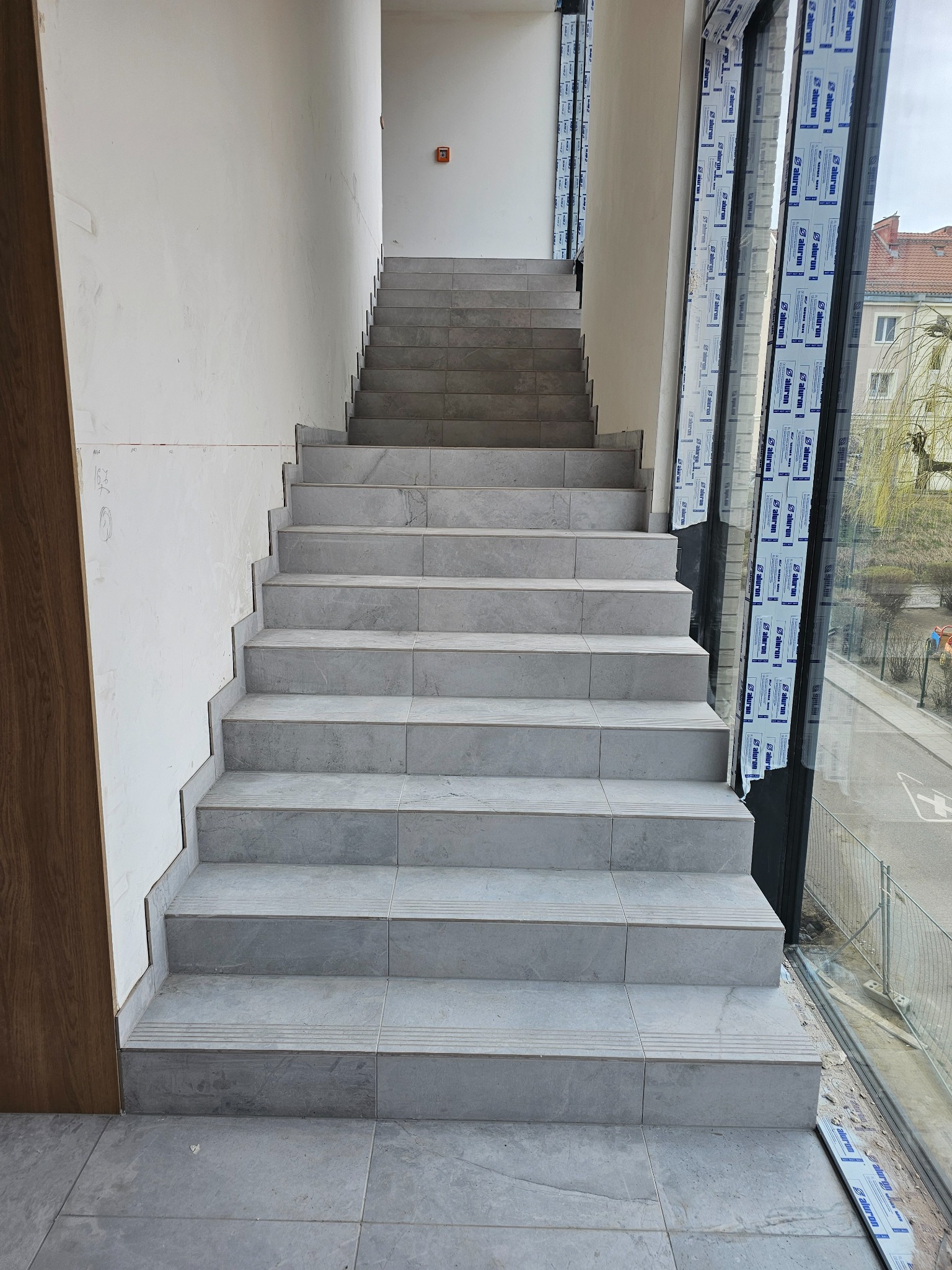 Wykończone schody wewnętrzne z szarych płytek, widok z dołu. Płytki ułożone równo, fuga czysta. Duże okno z widokiem na zewnątrz. Ściany przygotowane do malowania.