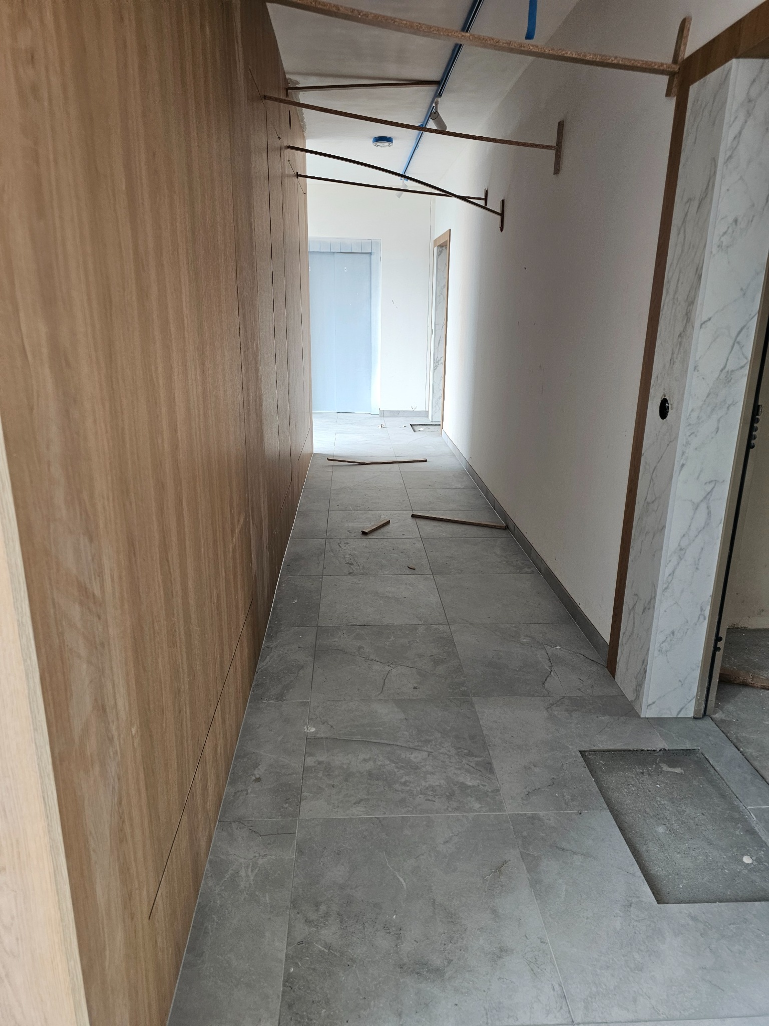 Szare płytki podłogowe w korytarzu, częściowo ułożone, z widocznym fragmentem podłogi bez płytek. W tle białe ściany i drzwi. Widoczne elementy konstrukcyjne sufitu.