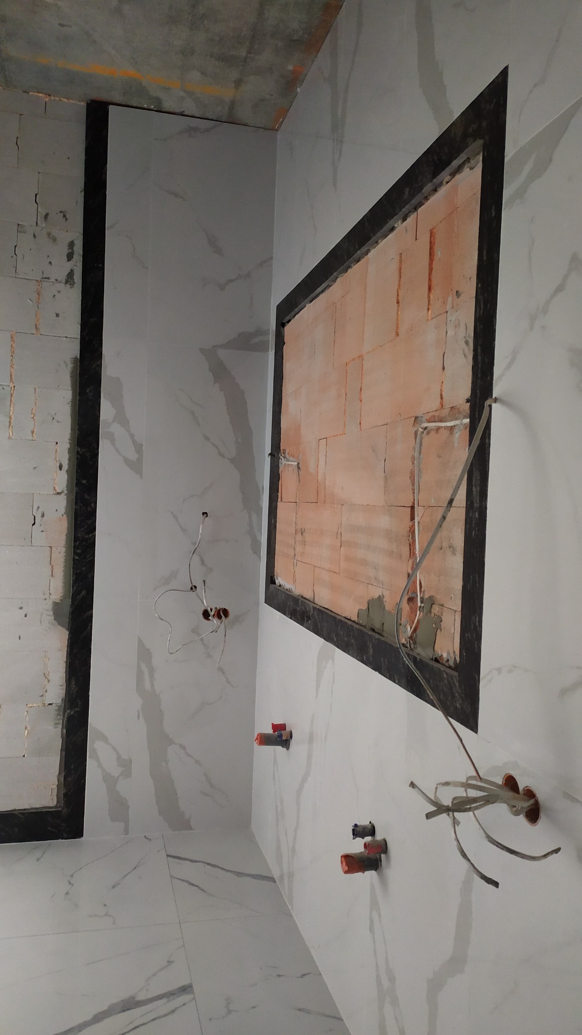 Wnętrze łazienki w trakcie wykańczania: białe płytki imitujące marmur na ścianach i podłodze, widoczne instalacje wodne i elektryczne, fragment ściany z cegły obramowany czarną ramą.