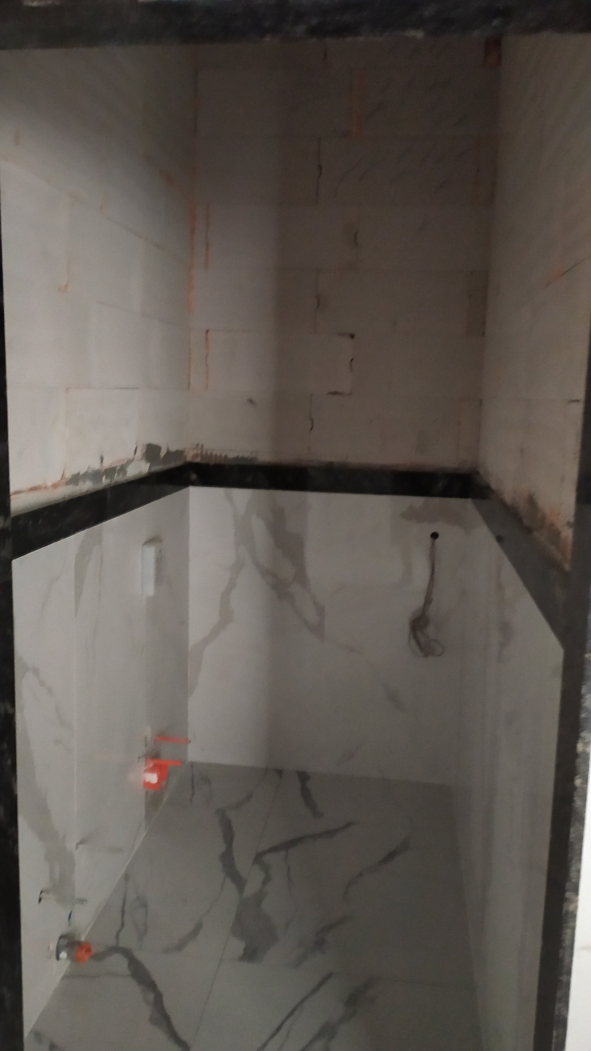 Wykończona wnęka z białymi płytkami imitującymi marmur na ścianach i podłodze. Widoczne czarne wykończenia i elementy instalacji. Użyto poziomowania płytek.