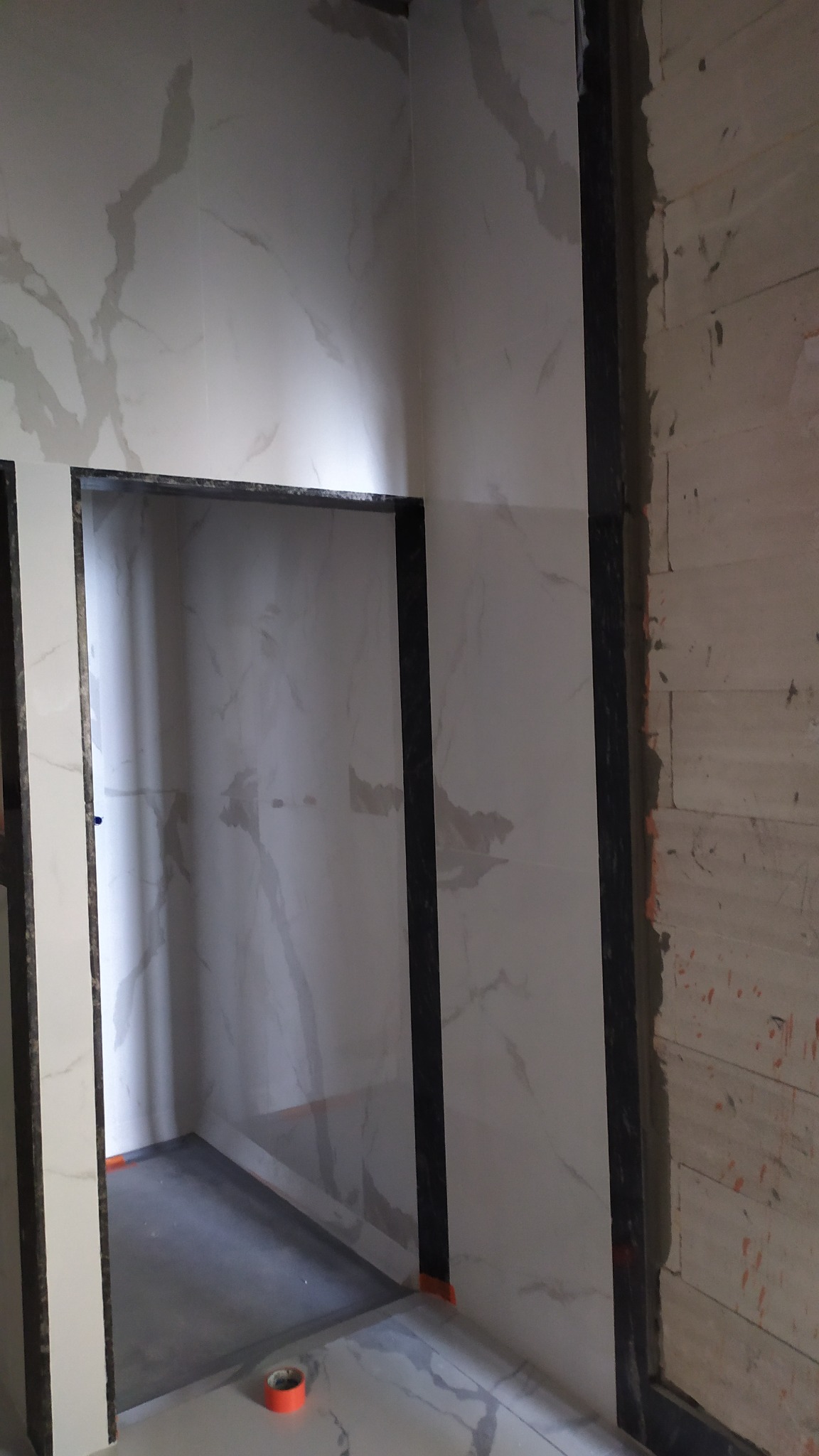 Wykończona ściana z płytek imitujących marmur w łazience, widoczny fragment kabiny prysznicowej z czarnym obramowaniem. Na podłodze taśma malarska i pomarańczowa rolka.