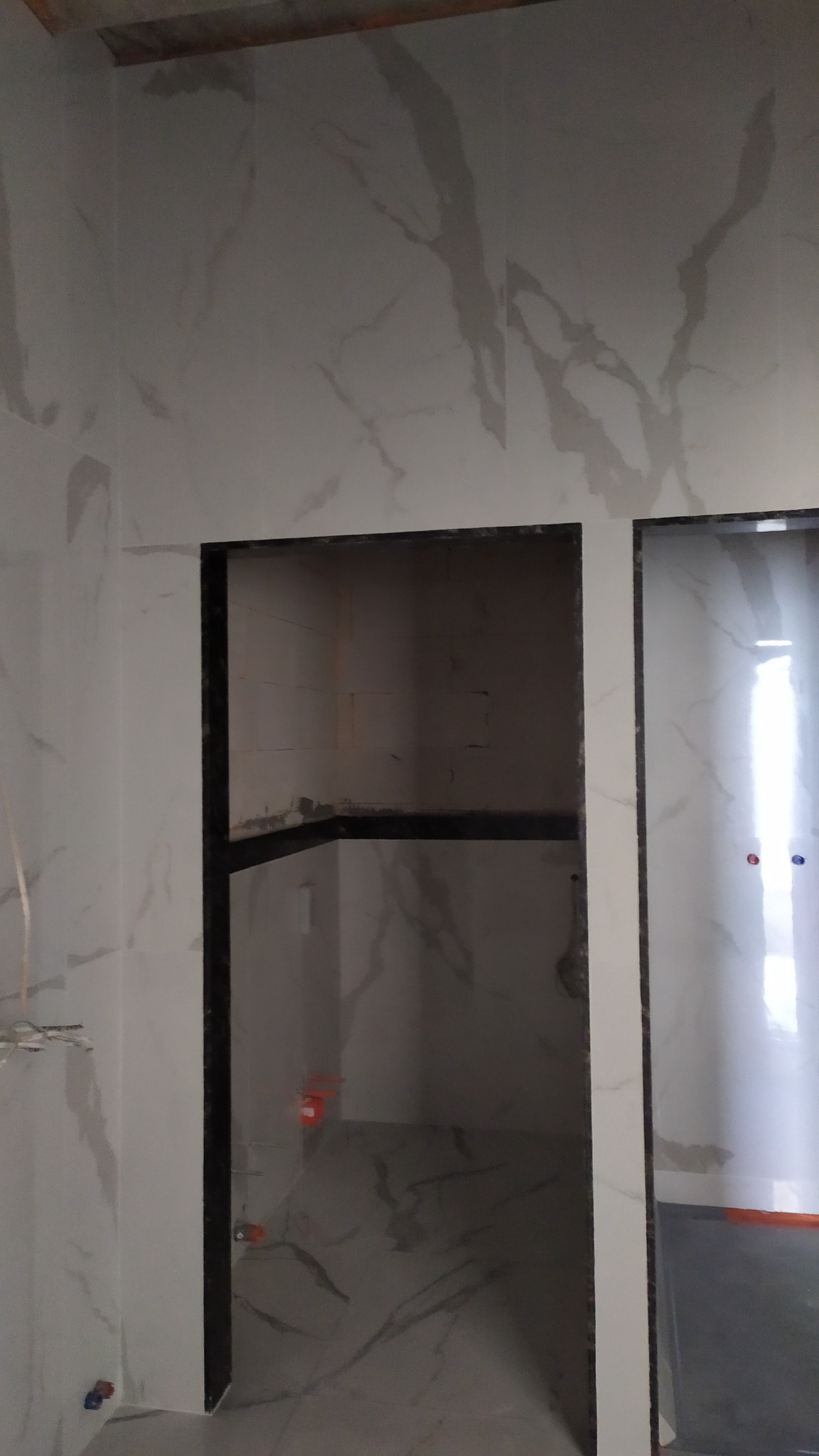 Wykończona wnęka z czarną ramą i fragment ściany z płytkami imitującymi marmur. Widoczny fragment instalacji wodnej. Minimalistyczny design łazienki w trakcie prac wykończeniowych.