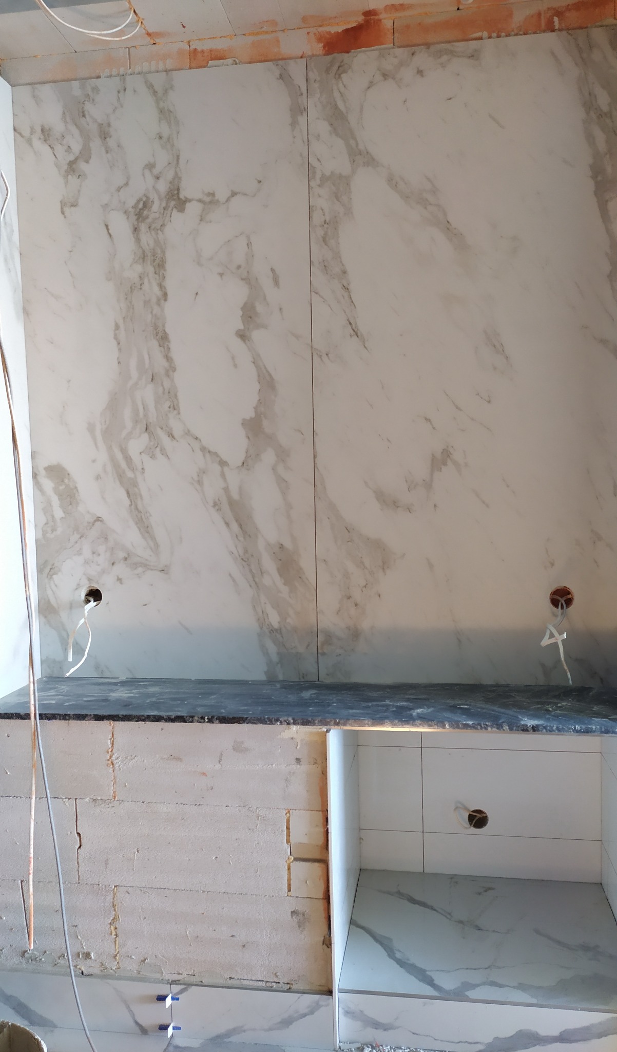 Ściana wyłożona płytkami imitującymi marmur, z widocznymi otworami na gniazdka. Częściowo wykończona zabudowa kuchenna z płytek. Widoczne elementy instalacji elektrycznej.