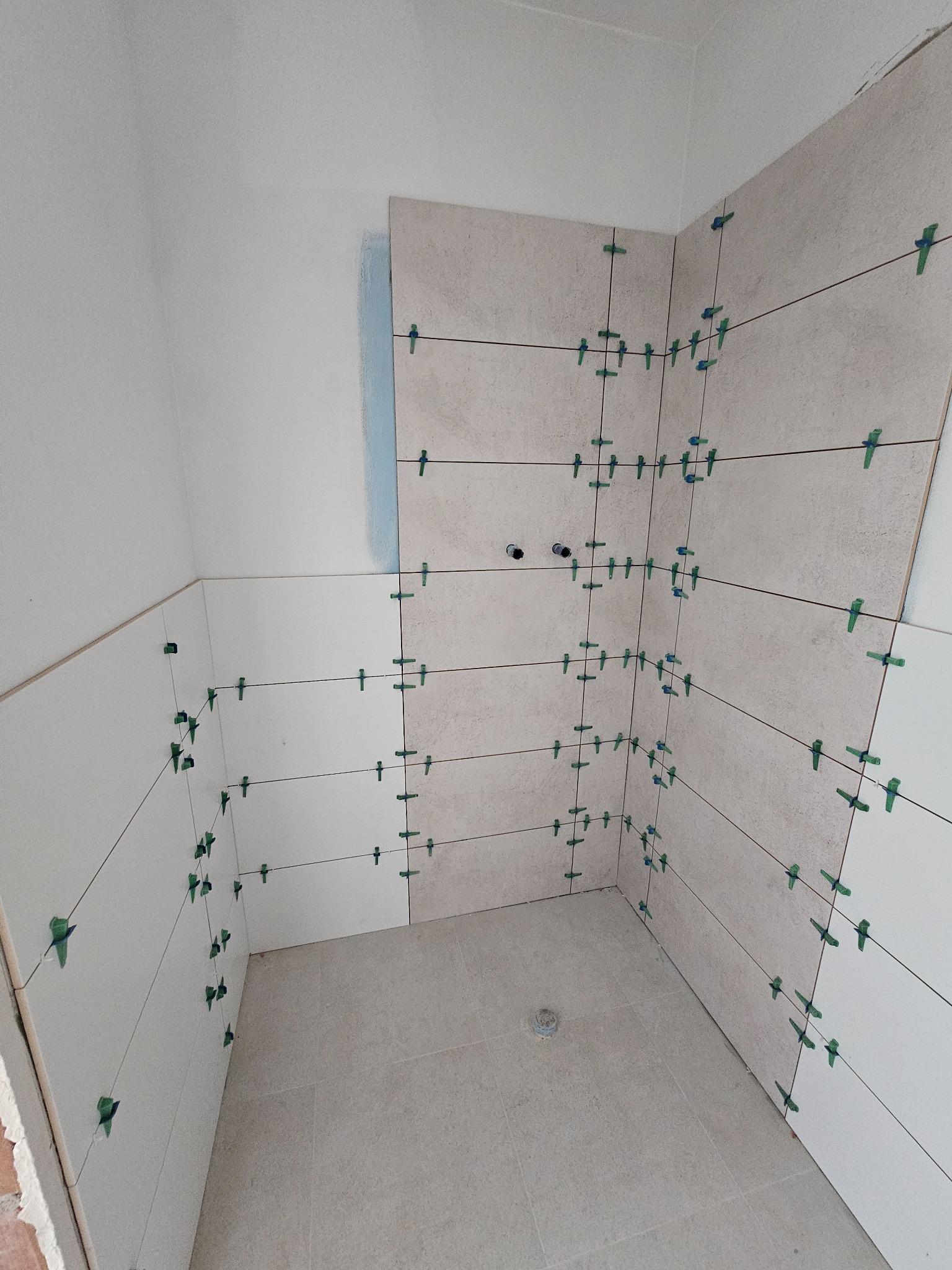 Ściana łazienki w trakcie układania płytek, z użyciem systemu poziomowania (klipsy i kliny). Widoczne fugi i otwory na instalacje. Częściowo ułożone płytki w odcieniach bieli i beżu.