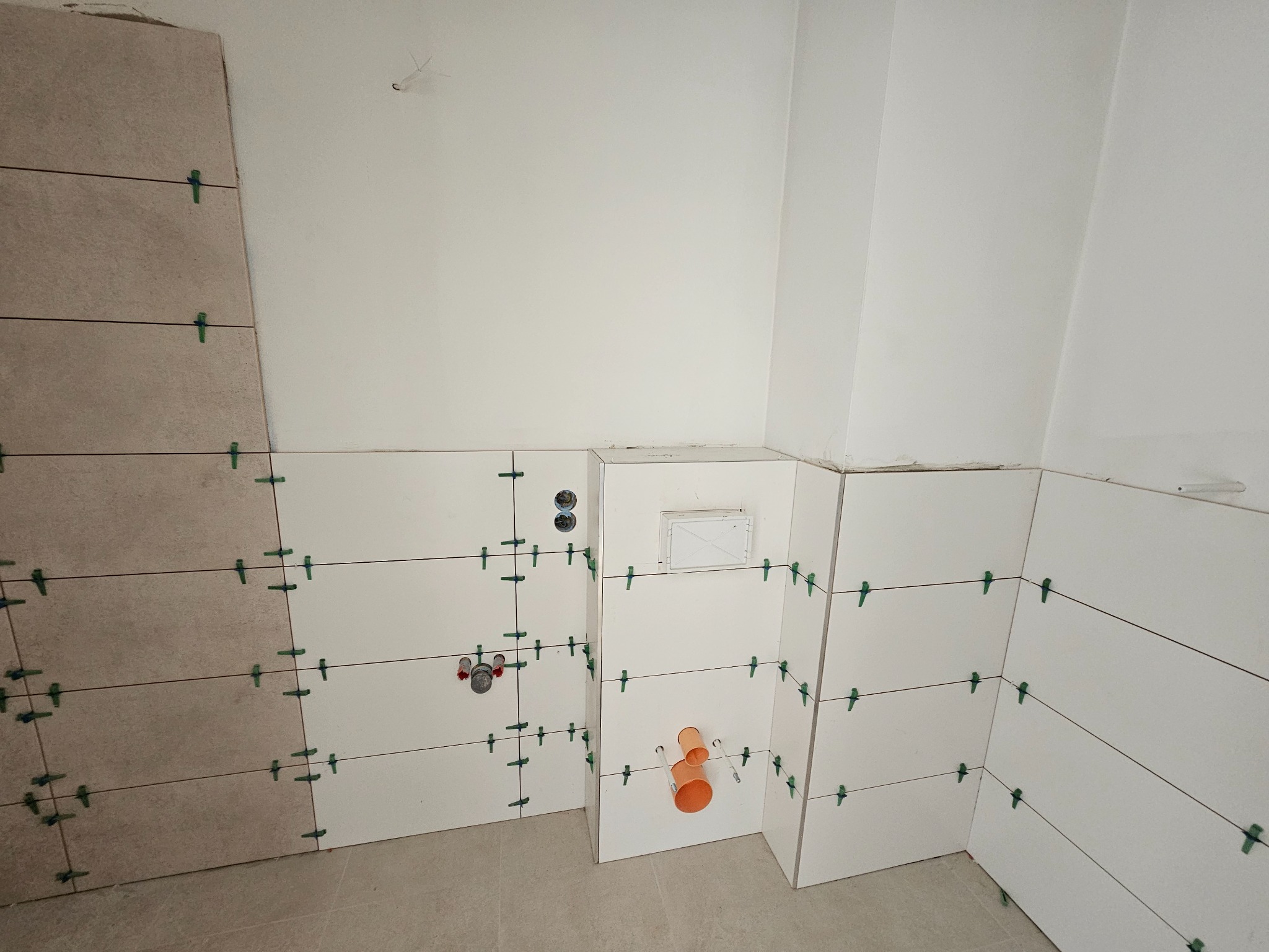 Układanie płytek ściennych w łazience z użyciem krzyżyków dystansowych. Widoczne instalacje wodno-kanalizacyjne i fragmenty płytek w odcieniach bieli i beżu.