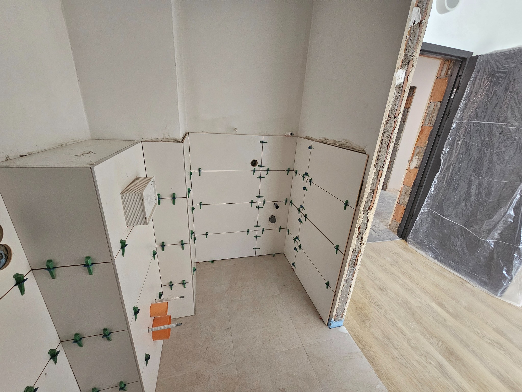 Białe płytki ścienne z zielonymi krzyżykami dystansowymi w trakcie montażu w pomieszczeniu. Widoczne fragmenty instalacji i podłoga z płytek.