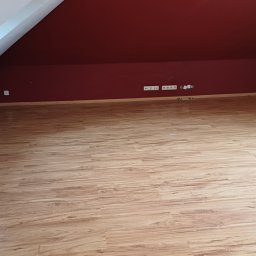 SHARK CLEANING - Poddasze z panelami podłogowymi w odcieniach drewna, bordowe ściany, biały grzejnik i skosy. Widoczne gniazdka elektryczne i listwy przypodłogowe.