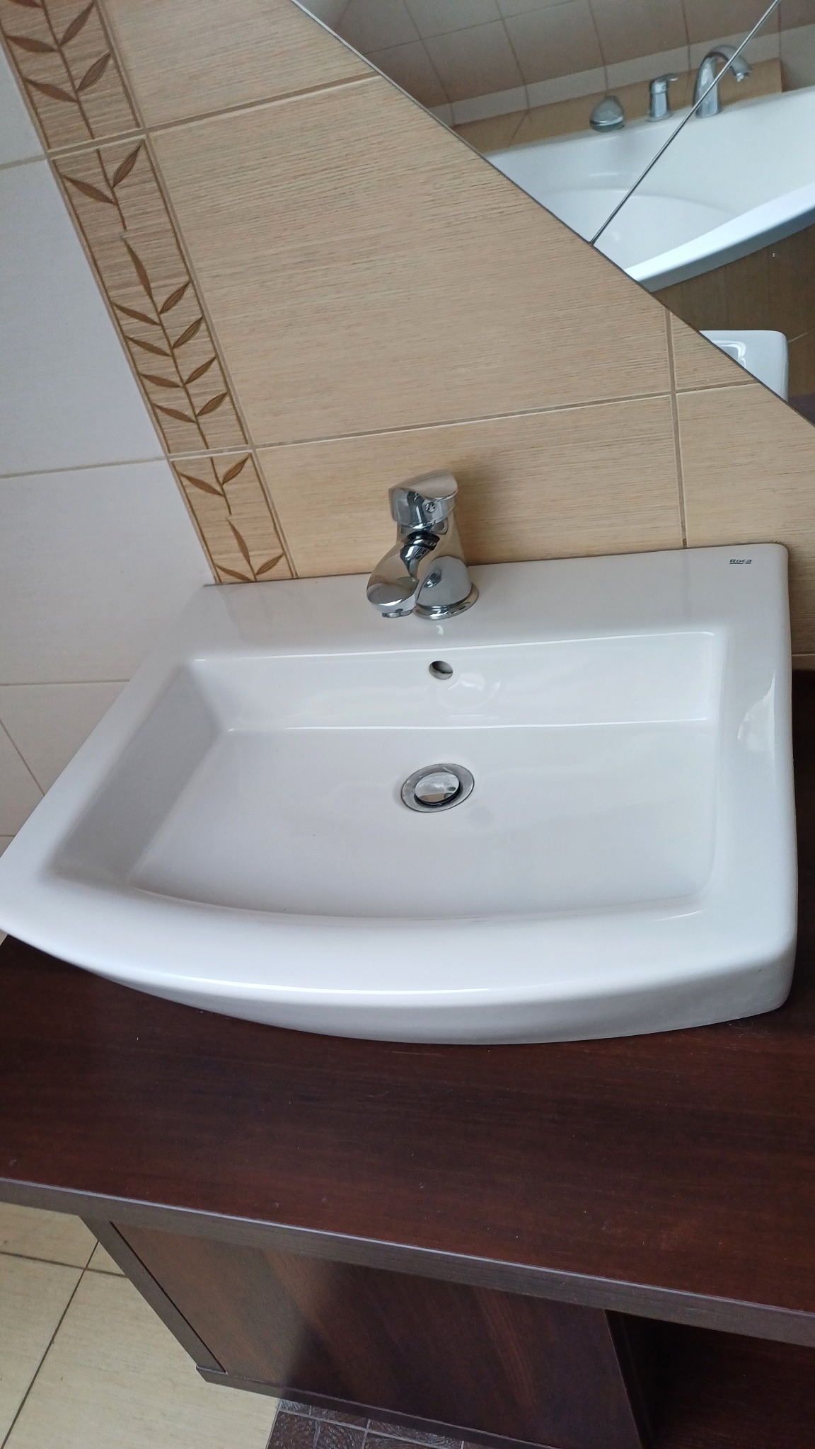 Biała umywalka Roca z chromowaną baterią na tle beżowych płytek z motywem roślinnym i lustra. Umywalka osadzona na ciemnobrązowej szafce.