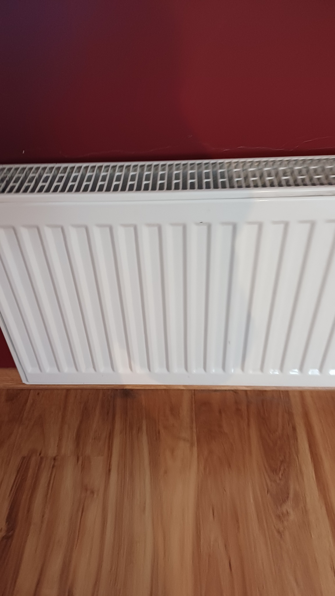 Biały grzejnik panelowy na tle bordowej ściany i drewnianej podłogi. Widok z góry, ukazujący czystość i estetykę po usłudze sprzątania.