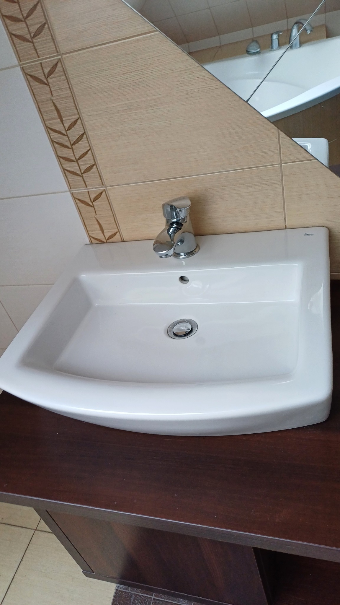 Biała umywalka Roca z chromowaną baterią na tle beżowych płytek z motywem liści, odbicie w lustrze ukazuje wannę. Czysta, gotowa do użytku.