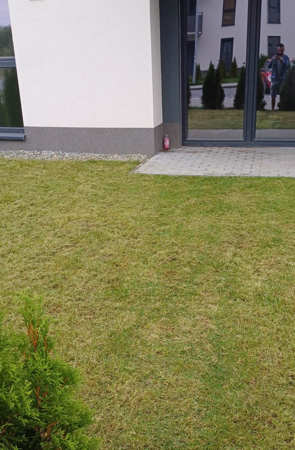 Nisko skoszony trawnik przy nowoczesnym domu z szarym cokołem i dużymi oknami. W odbiciu okna widoczna osoba robiąca zdjęcie.