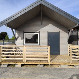 ENERGO DOM SPÓŁKA Z OGRANICZONĄ ODPOWIEDZIALNOŚCIĄ - Gotowy, szary dom szkieletowy z drewnianym tarasem w Jagniewicach. Widoczne materiały budowlane w tle. Dach dwuspadowy, okno z białą ramą, szare drzwi.