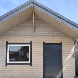 ENERGO DOM SPÓŁKA Z OGRANICZONĄ ODPOWIEDZIALNOŚCIĄ - Elewacja domu szkieletowego z jasnego drewna z ciemnymi obramowaniami okna i drzwi. Widoczny fragment dachu i boczna ściana sąsiedniego budynku.