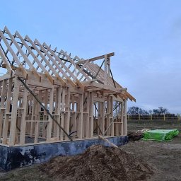 ENERGO DOM SPÓŁKA Z OGRANICZONĄ ODPOWIEDZIALNOŚCIĄ - Surowy szkielet domu w trakcie budowy, widoczna drewniana konstrukcja ścian i dachu na fundamencie, z ziemią i zielonym materiałem budowlanym w tle.